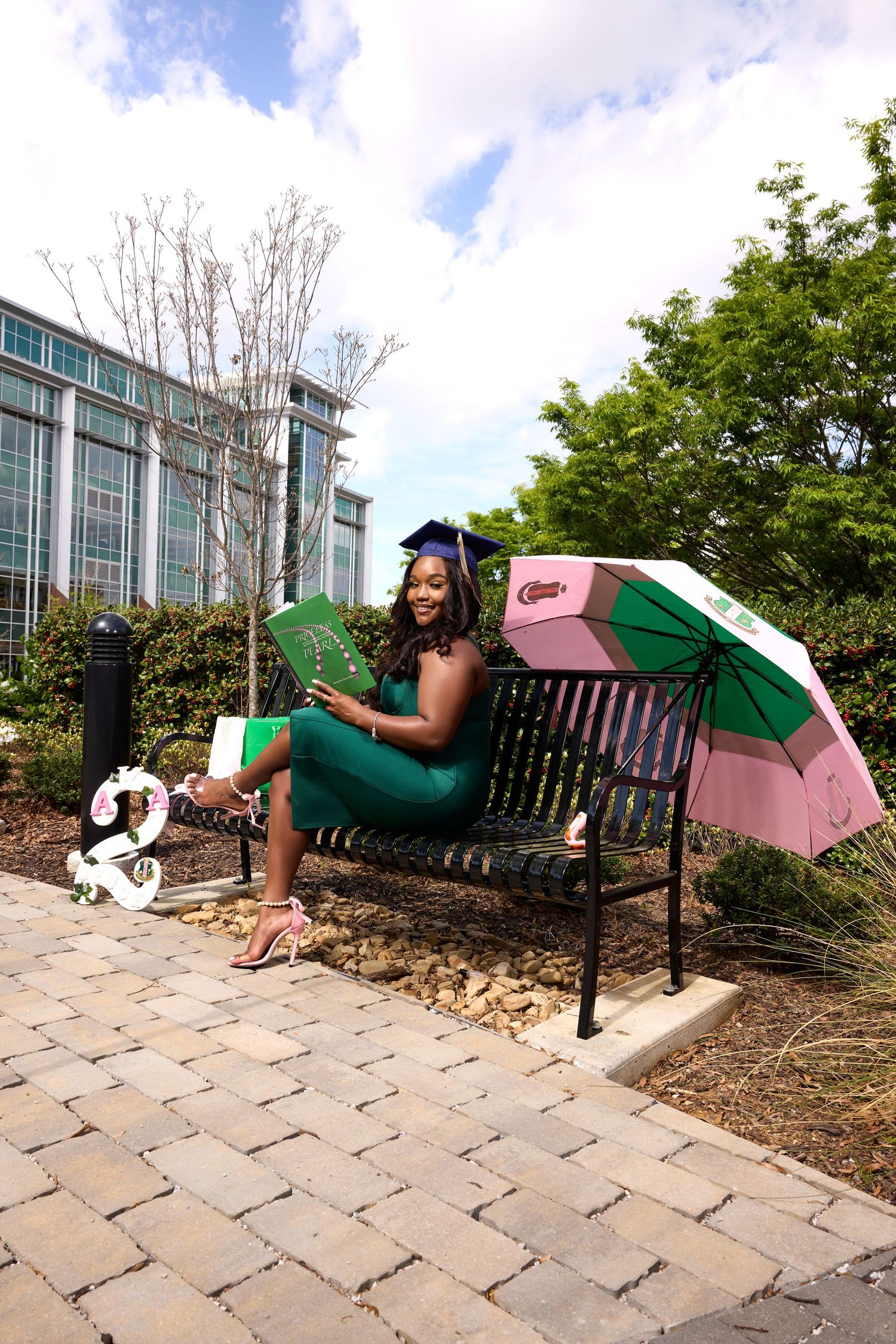 A graduate in green dress poses playfully on bench with pink and green umbrella outdoors.