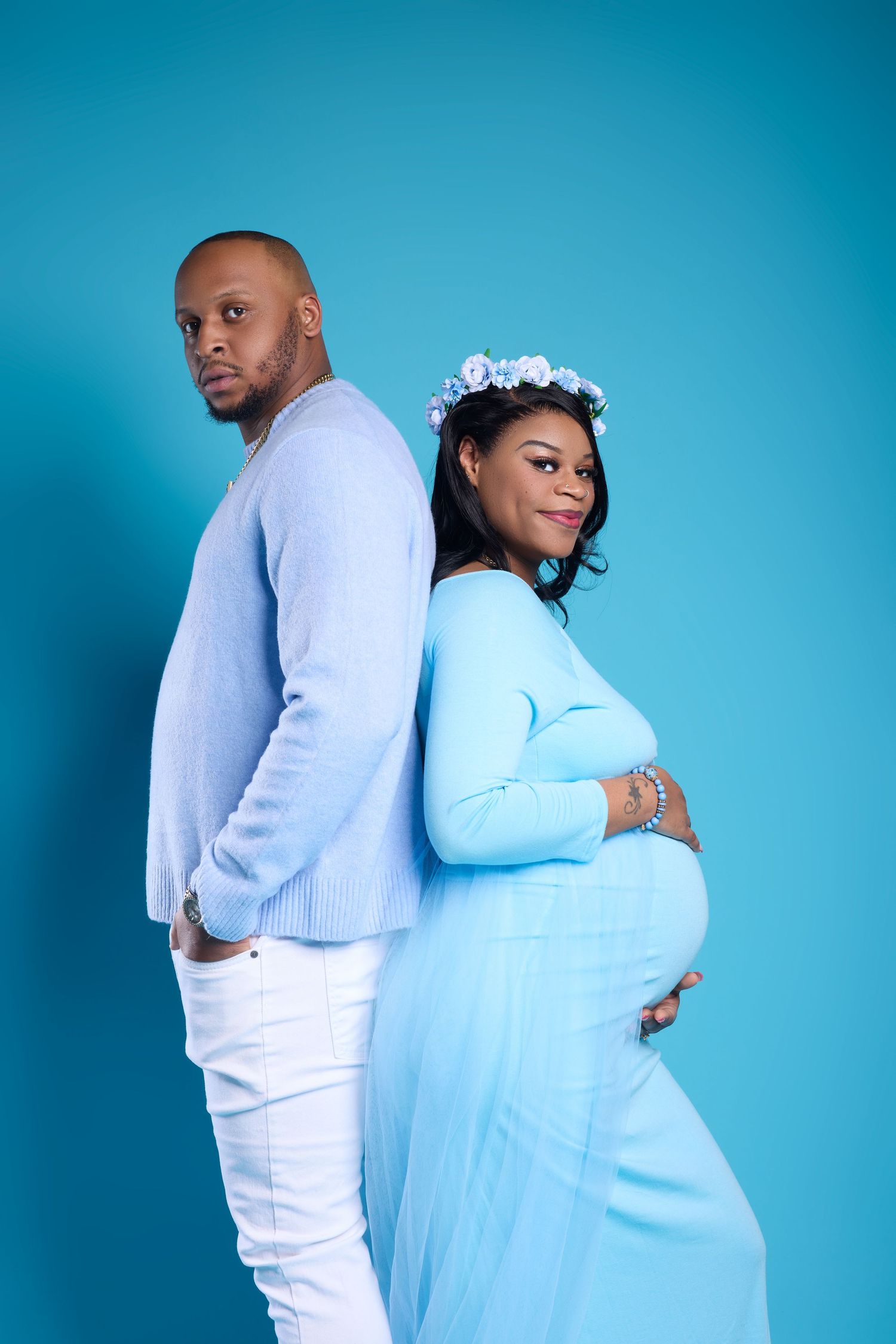 A pregnant woman in a blue gown and floral crown poses back-to-back with a man in a blue sweater against a blue backdrop.