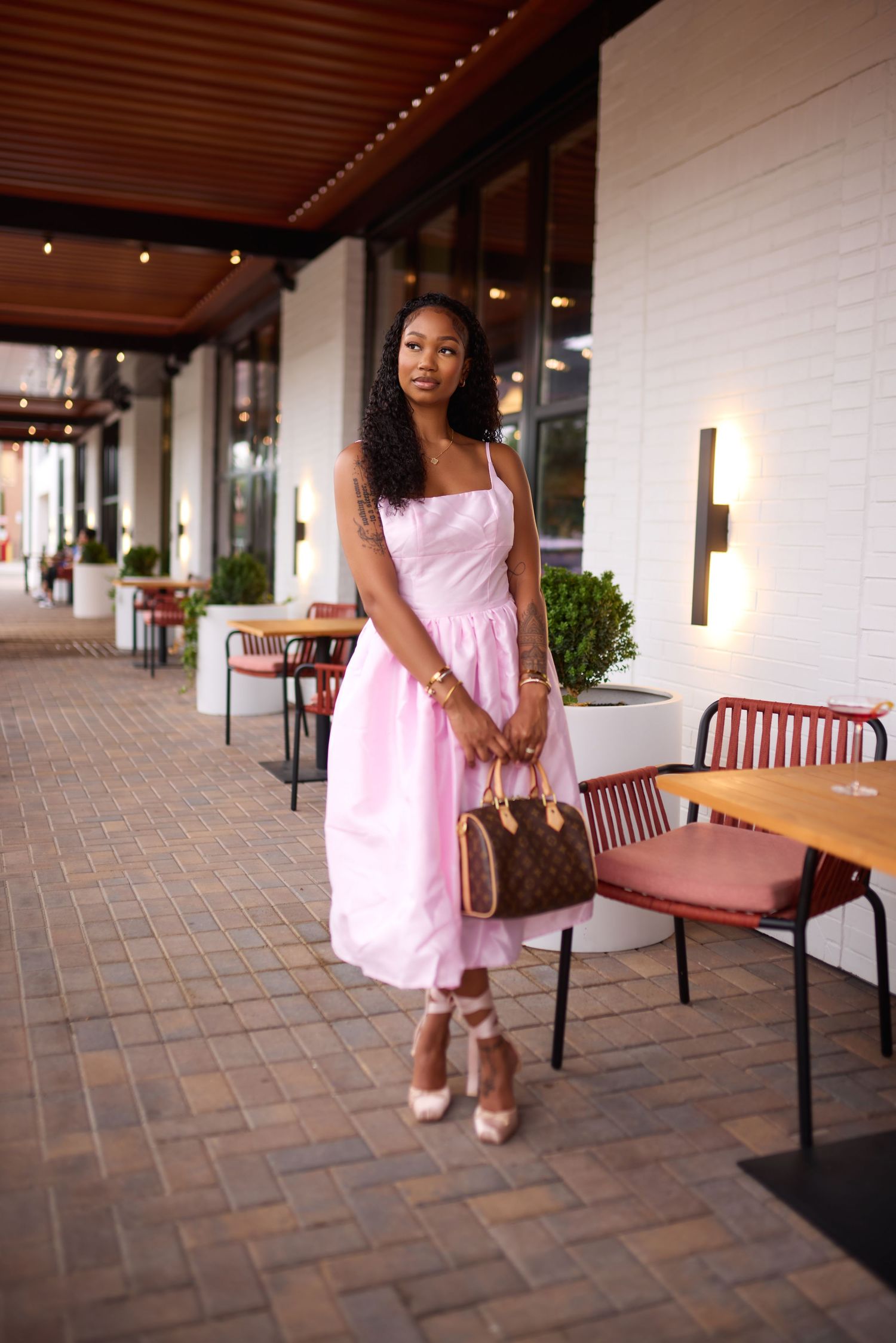 Woman in pink midi dress holding Louis Vuitton bag, wearing nude heels, posing at outdoor restaurant patio.