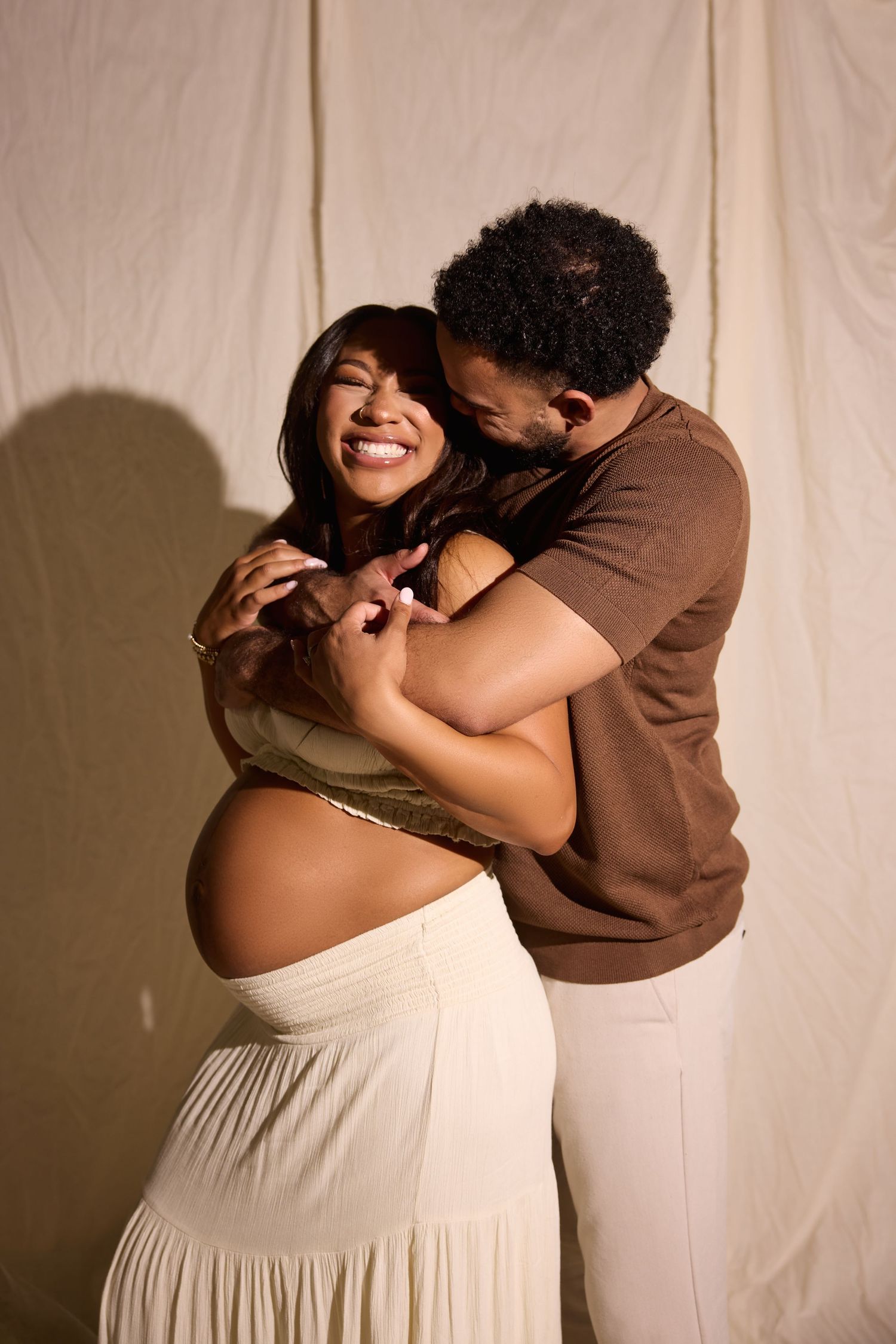 A joyful pregnant woman embraces her partner during a maternity photoshoot against a neutral fabric backdrop.
