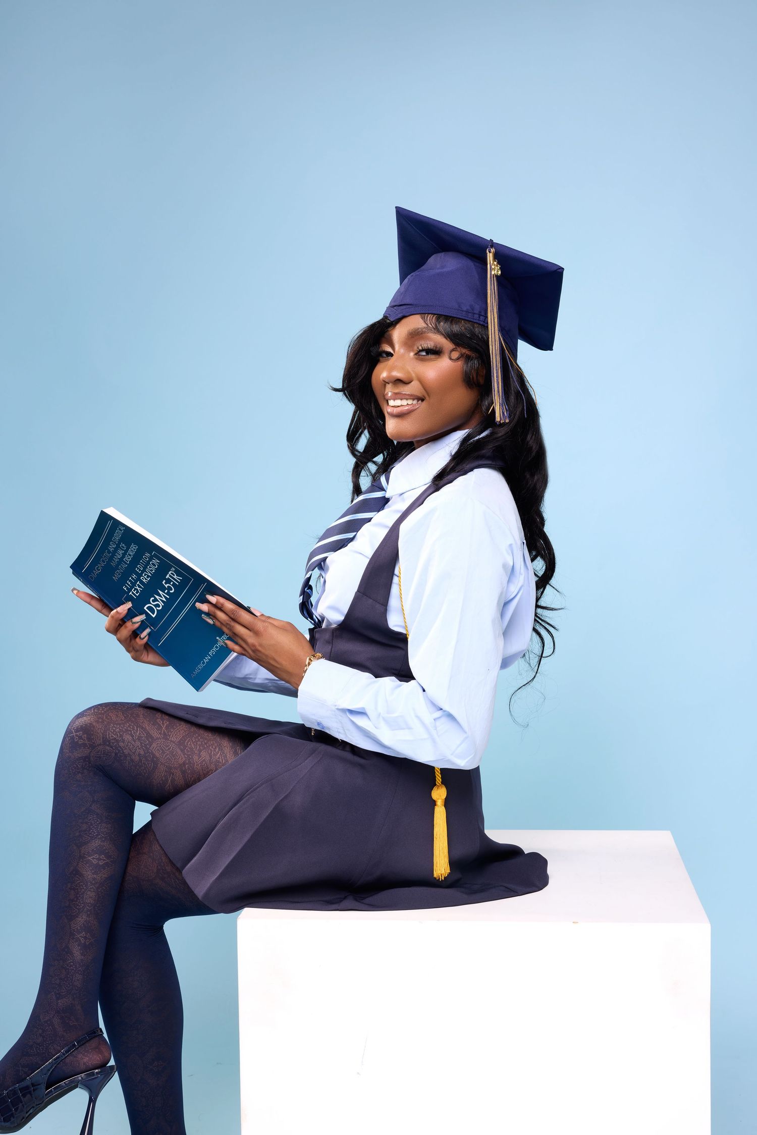 Graduate in cap and gown poses with books against light blue backdrop.