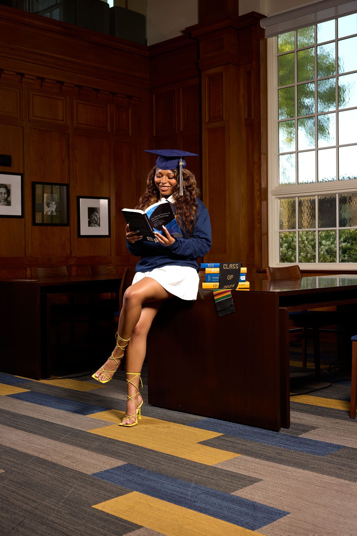 Graduate sits on wooden bench with books in traditional academic library setting.