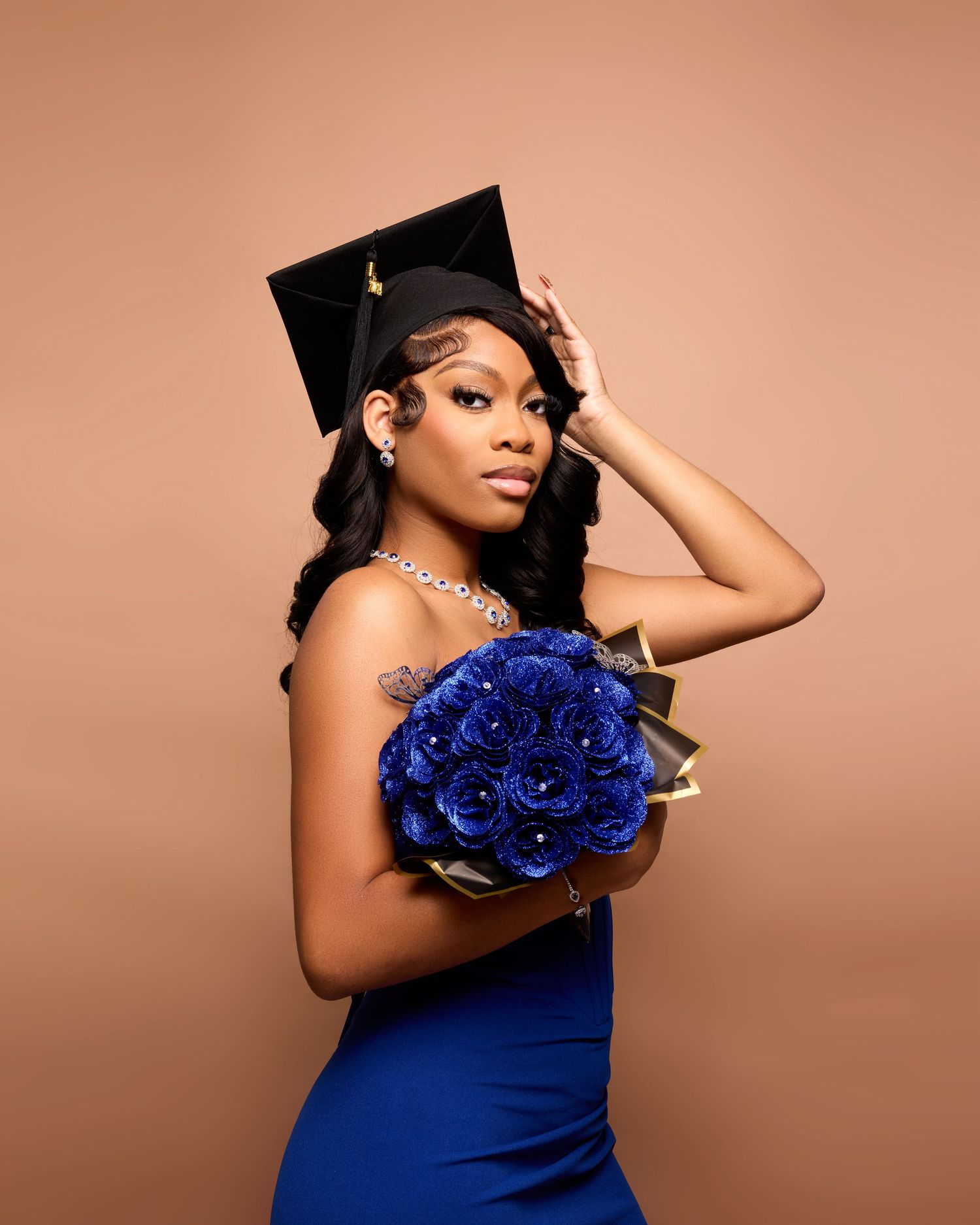 Graduate holding blue roses poses in cap and gown against neutral backdrop.