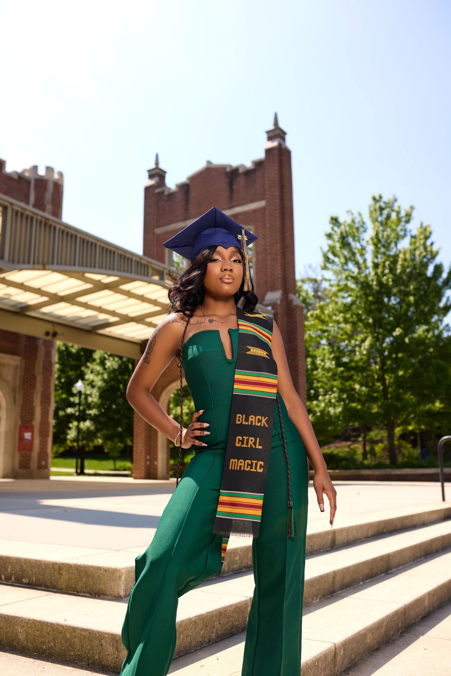 Graduate poses on concrete steps wearing green graduation gown with kente stole.