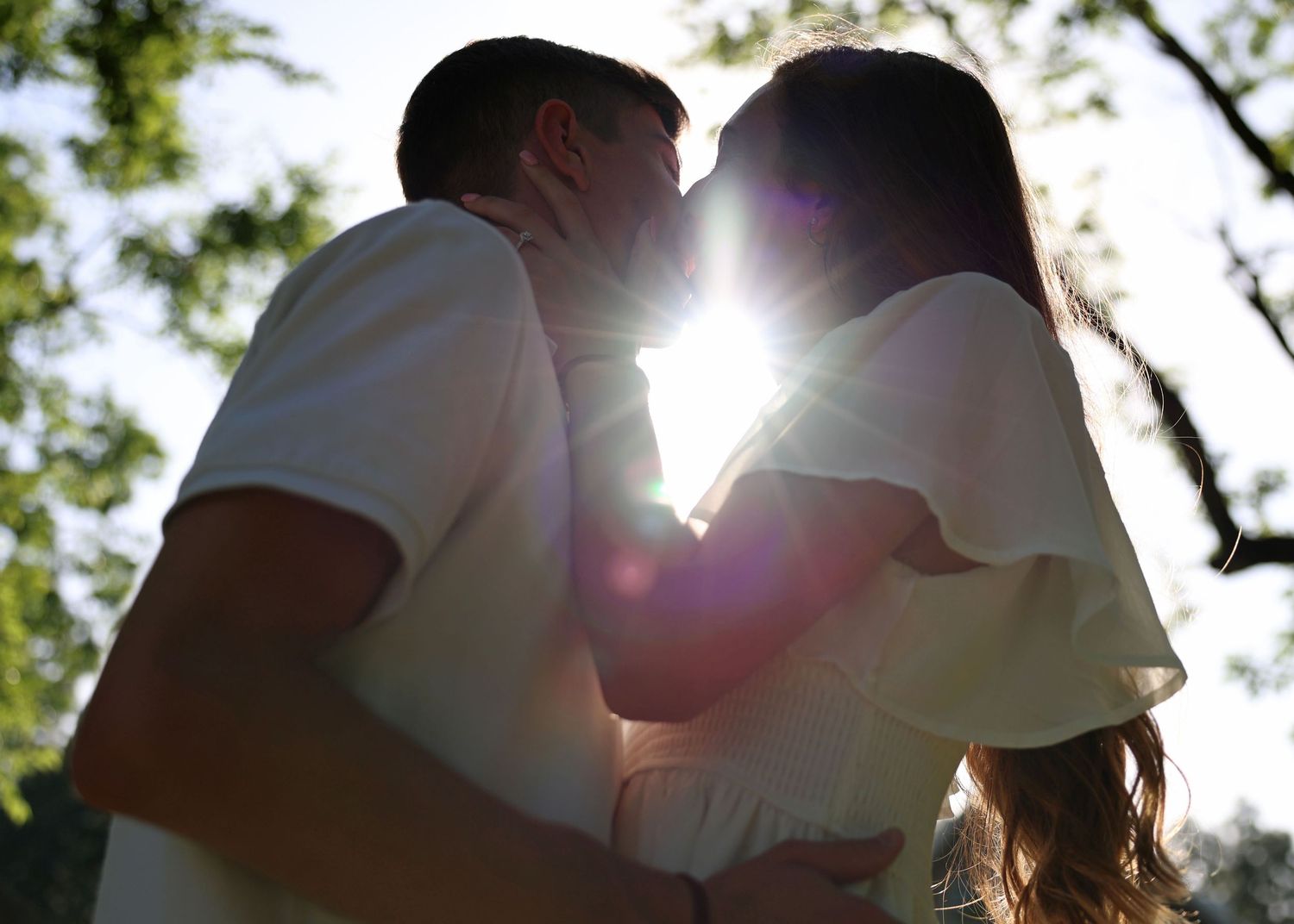 Silhouetted couple sharing an intimate moment against bright sunlight filtering through trees.
