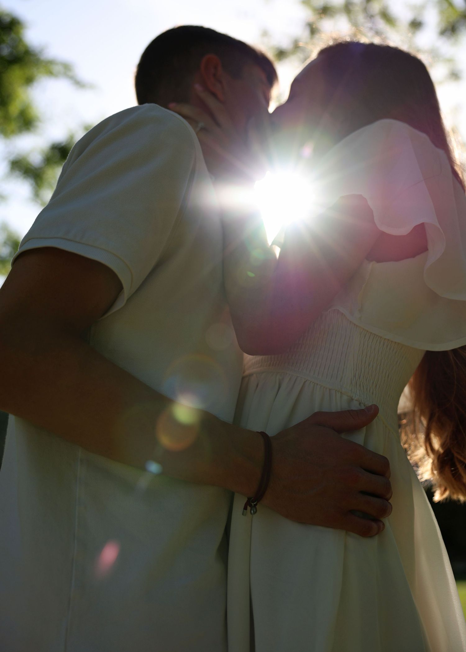 Silhouetted couple in white clothing embracing with sun flare between them.
