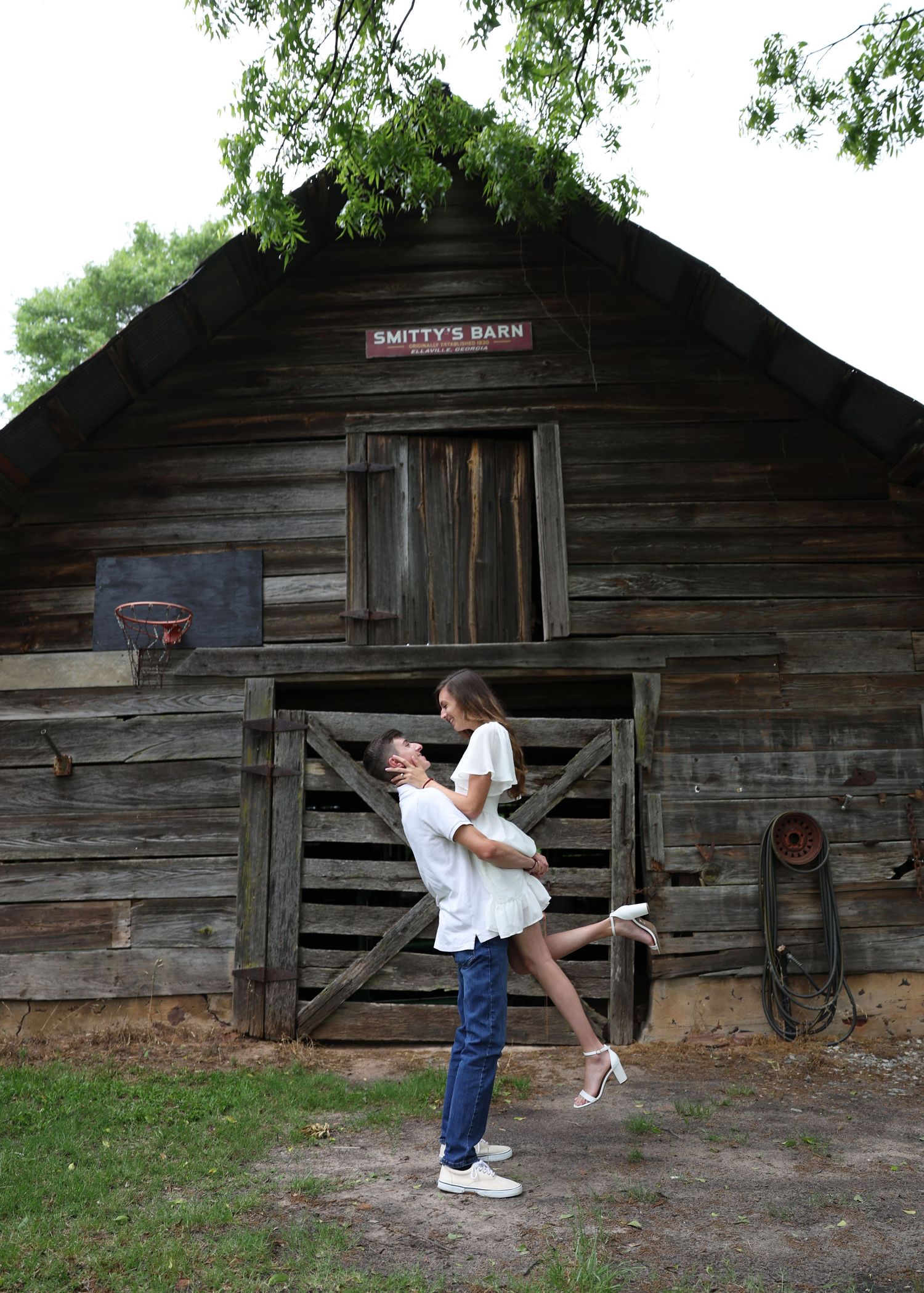 Couple in playful pose outside rustic wooden barn with 'Smith's Barn' signage.