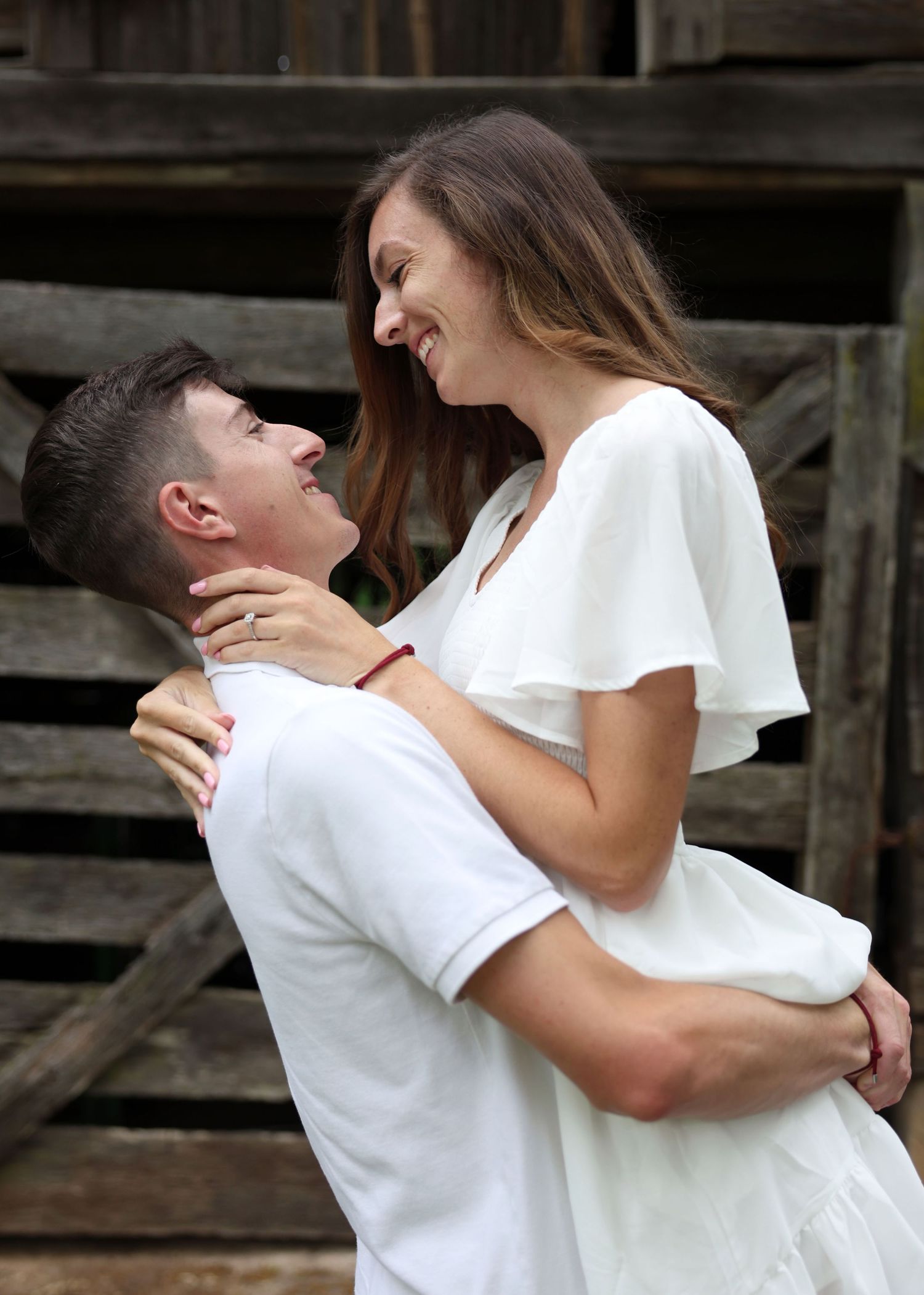 Romantic couple in white outfits sharing a joyful embrace against a rustic wooden backdrop.