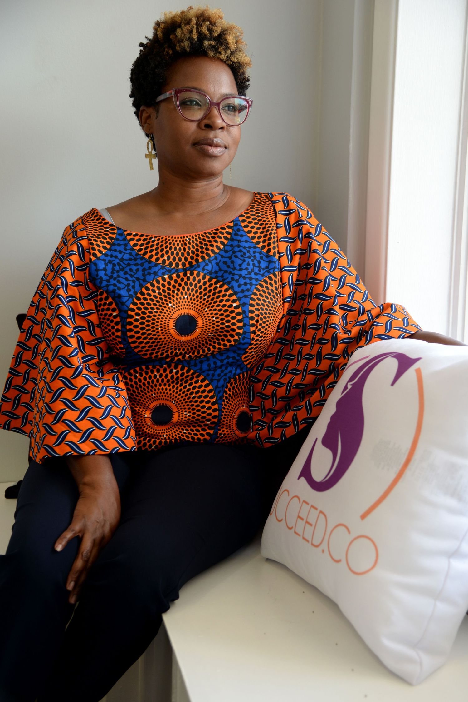 A person wearing glasses and a vibrant African print dress with orange and blue circular patterns sits beside a white pillow.
