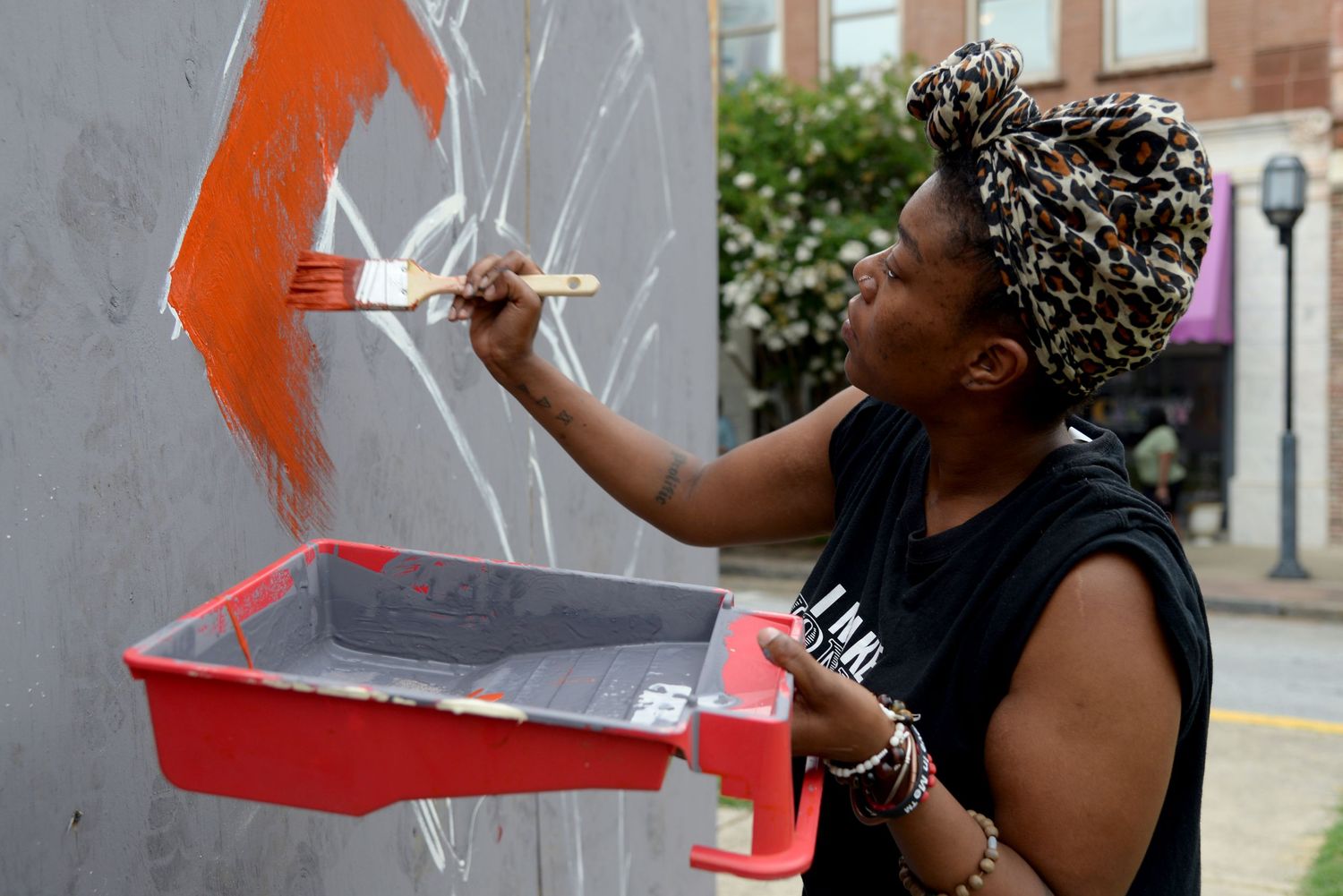 Person wearing a headwrap painting with orange paint on a wall outdoors.