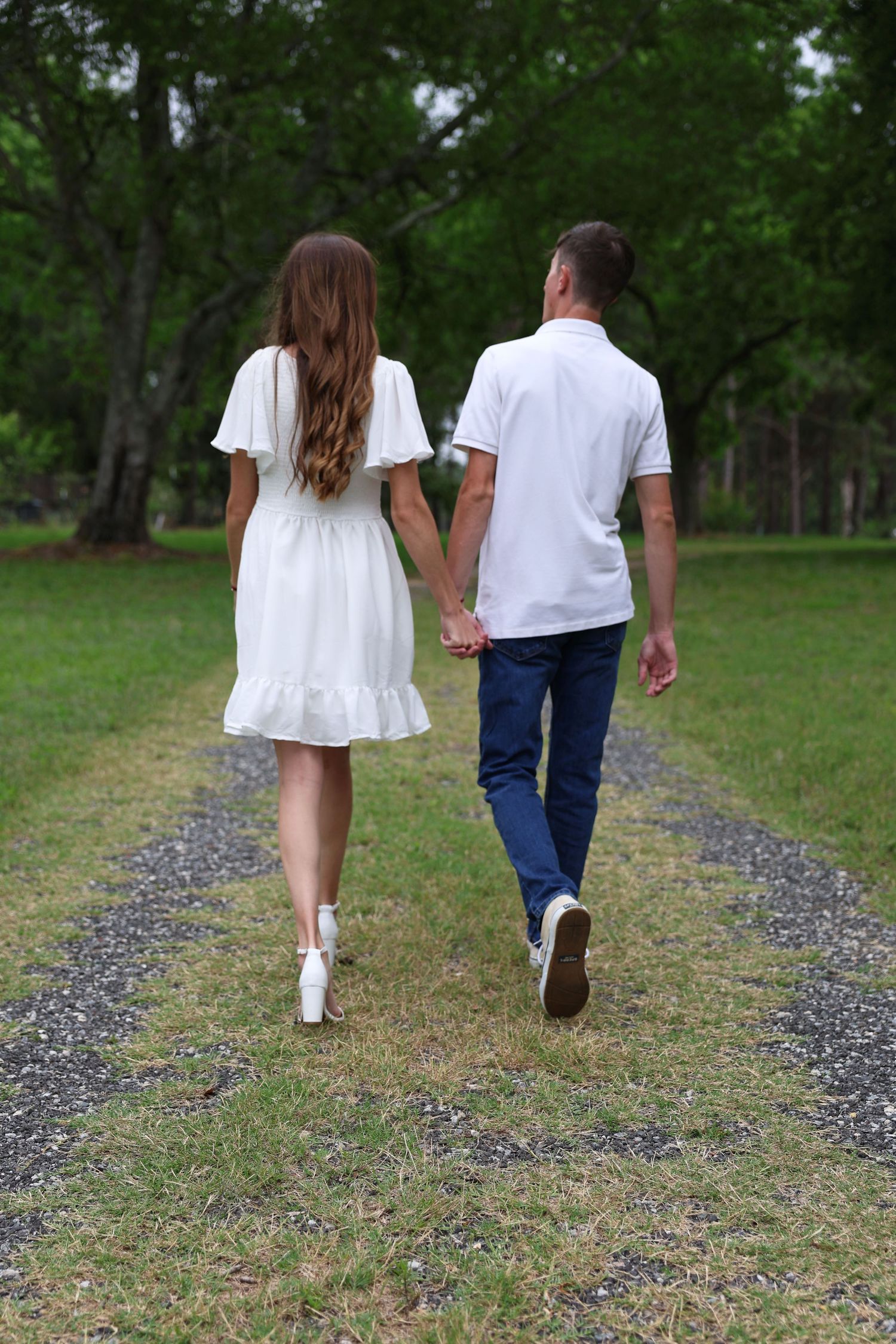 A couple in white outfits walking hand in hand down a gravel path in a lush green park.