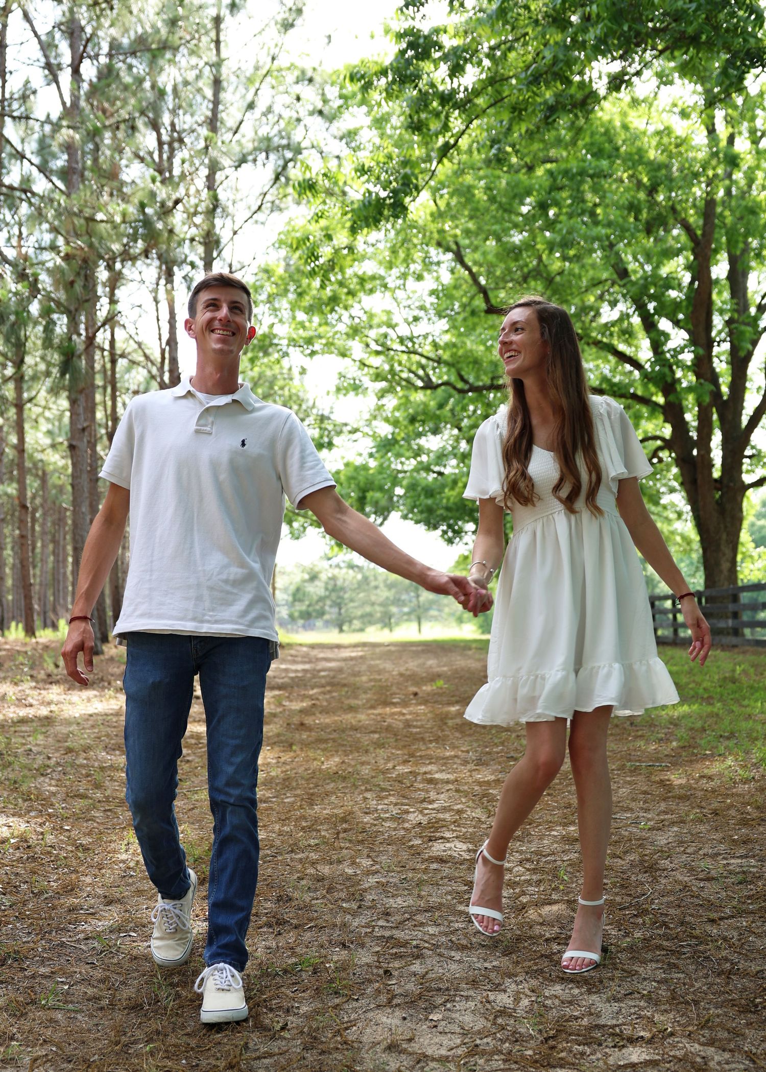 Couple in coordinated white outfits walking and laughing together on a wooded trail.