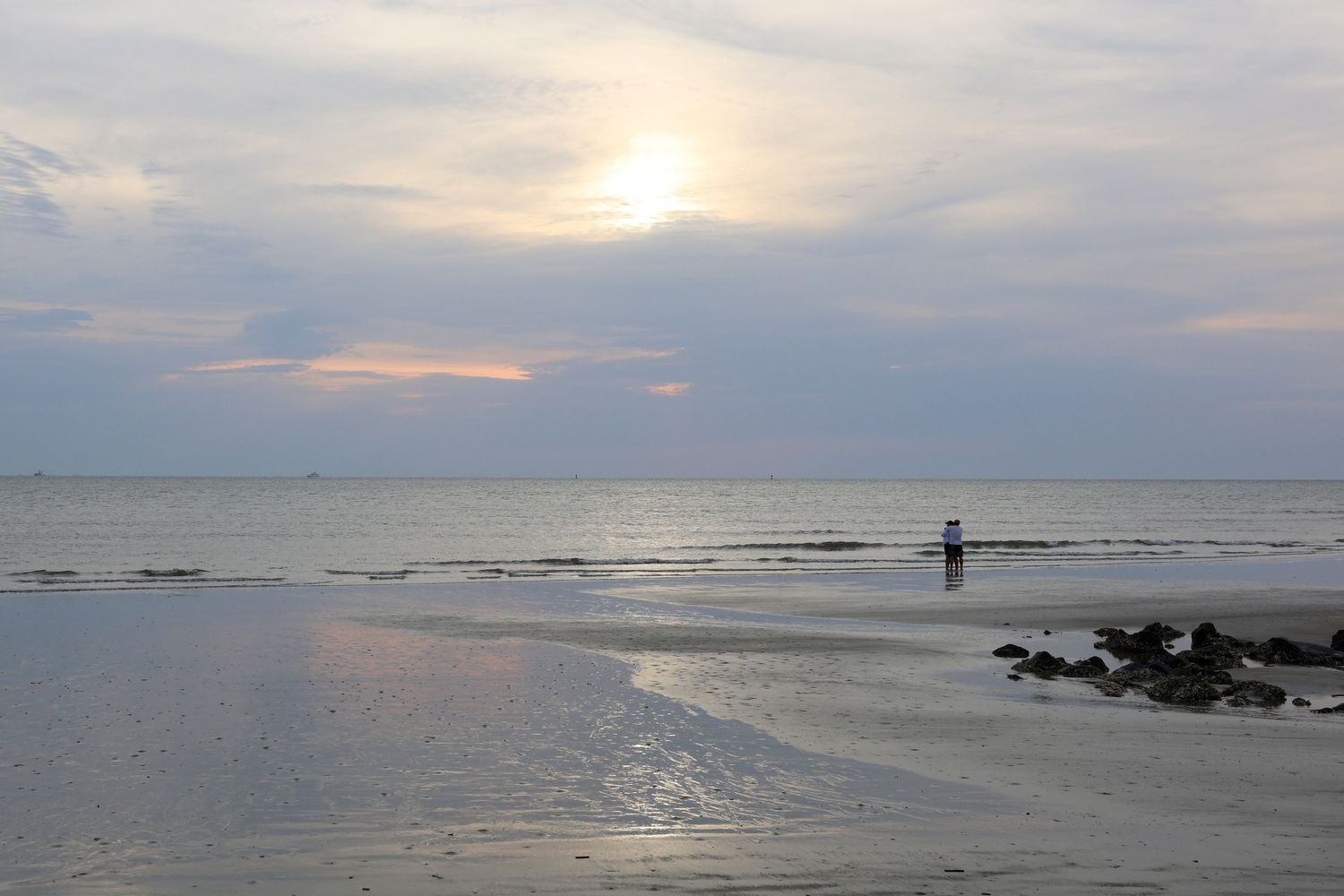 Solitary figure stands on a beach at sunset with cloudy skies and calm ocean waters.