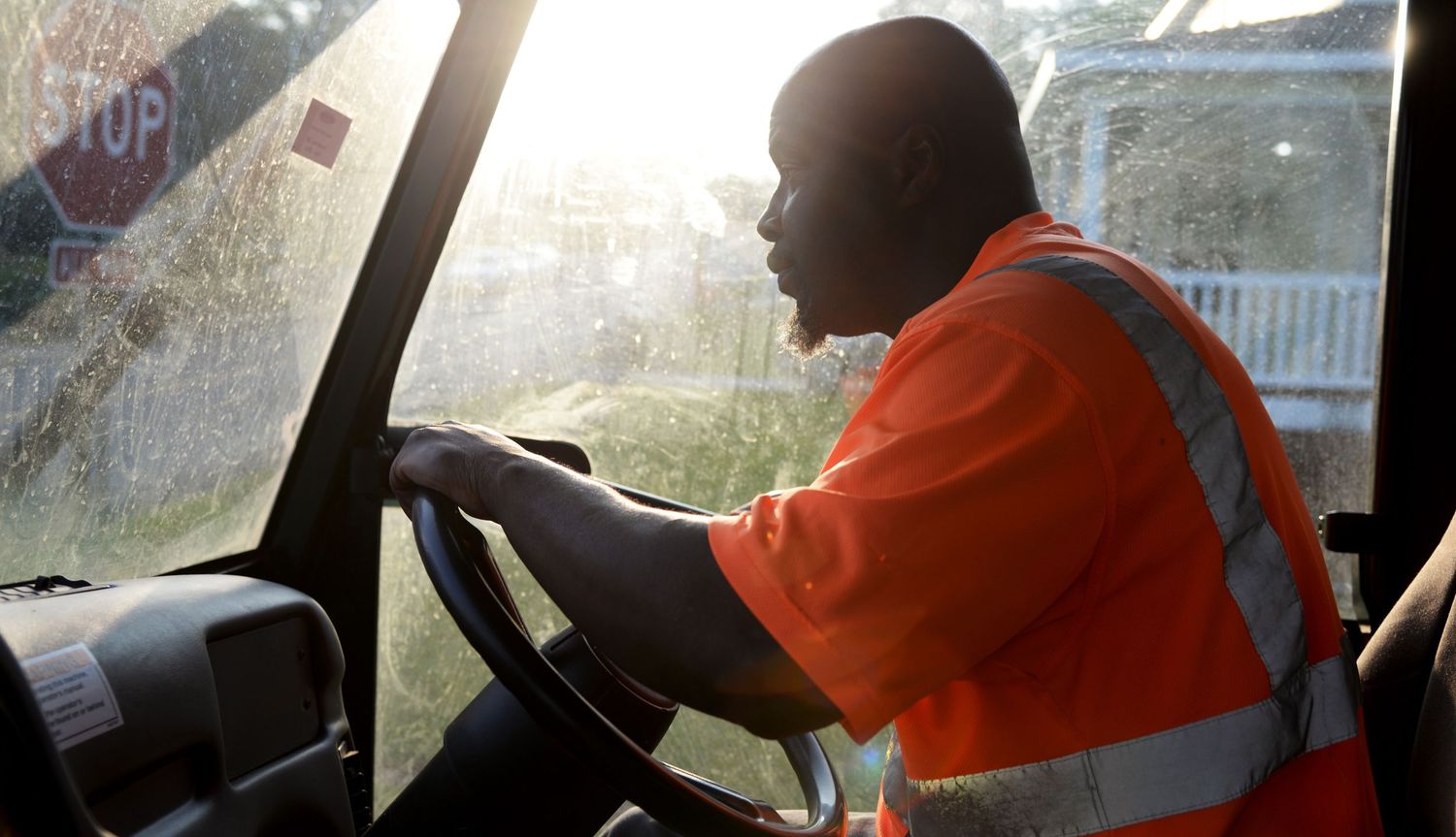 Worker in orange safety vest driving a vehicle during sunset.
