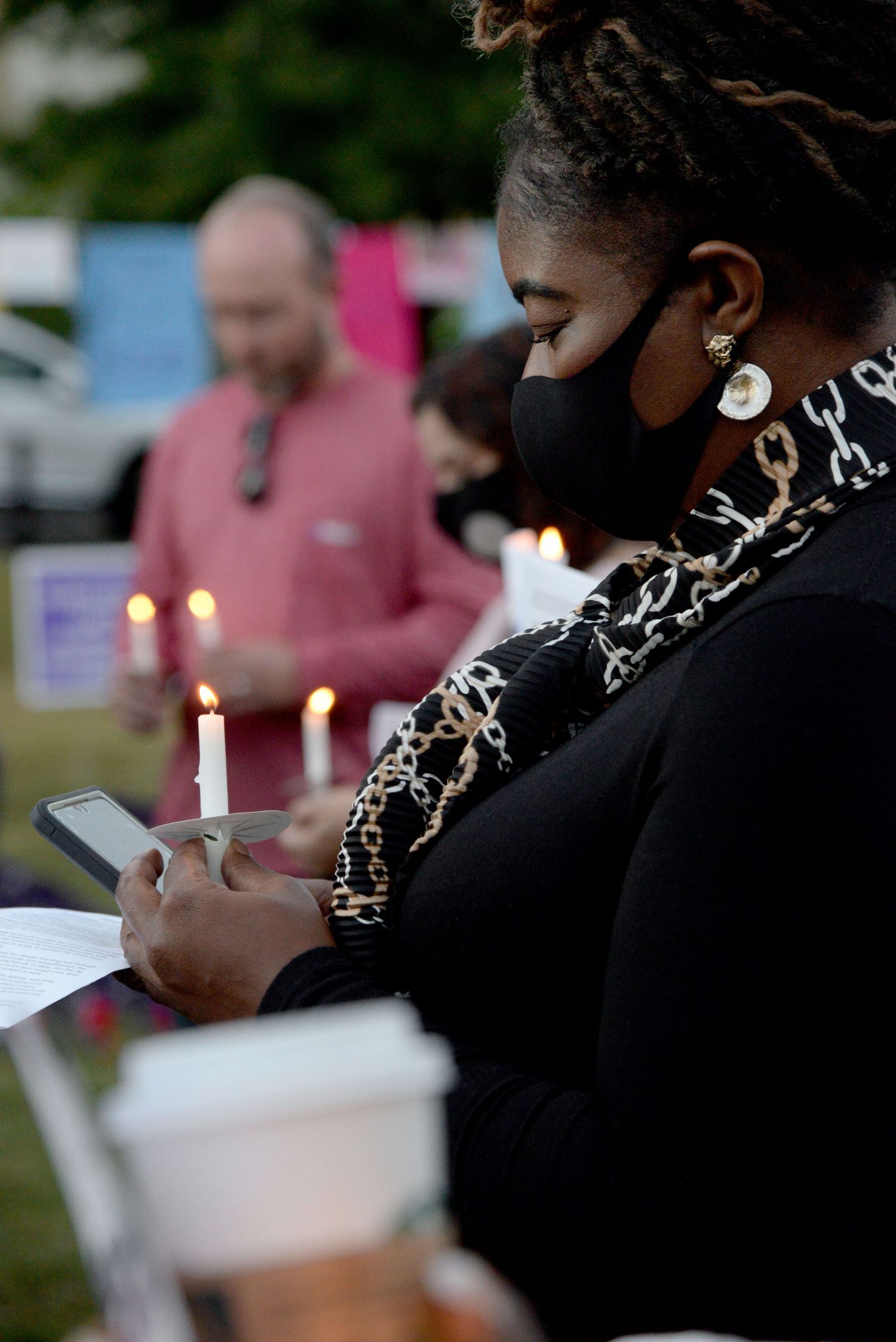 Person at vigil in black clothing and face mask holding a candle while checking their phone at night.