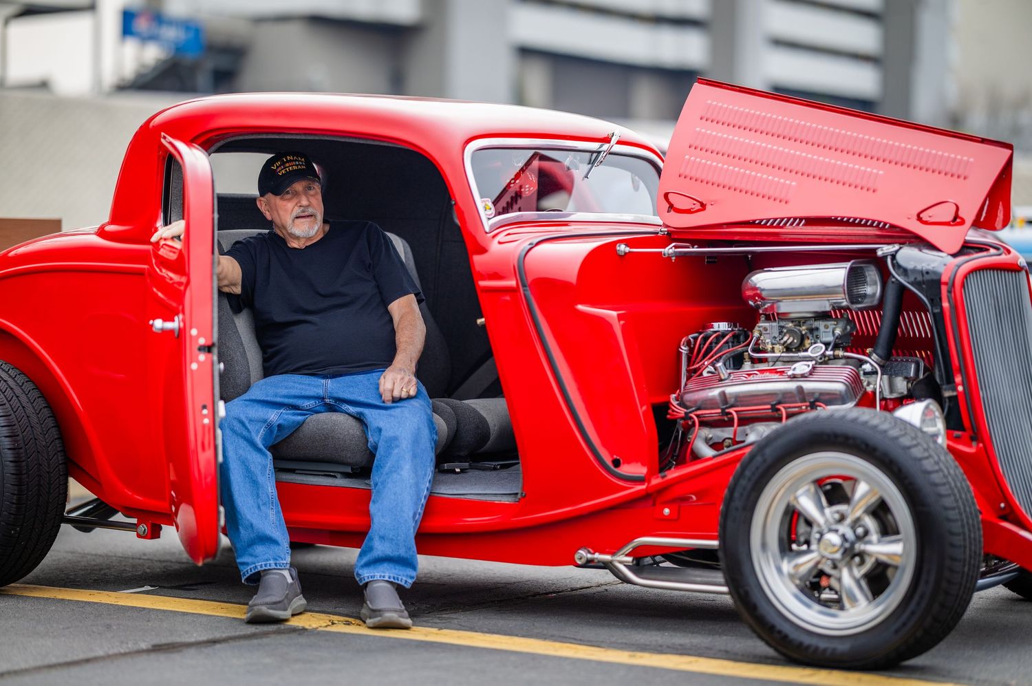 Man sitting in a classic red hot rod car with chrome engine details at an outdoor car show.