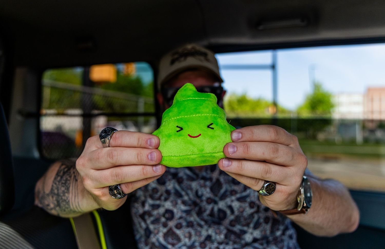 Tattooed person holds a small green smiling frog plush toy inside a vehicle with trees visible outside.