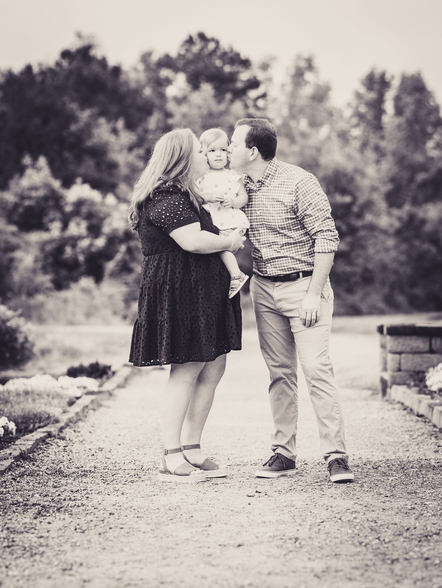 Black and white family portrait of parents kissing their toddler on a garden path surrounded by lush trees.
