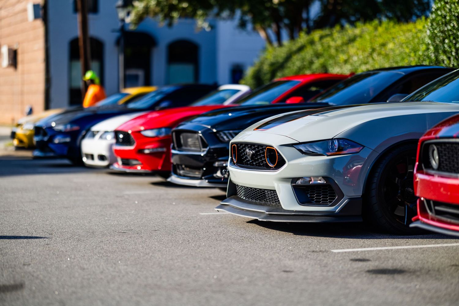 Row of colorful Ford Mustangs parked in a sunny lot, showcasing red, white, and blue sports cars at a car meet.