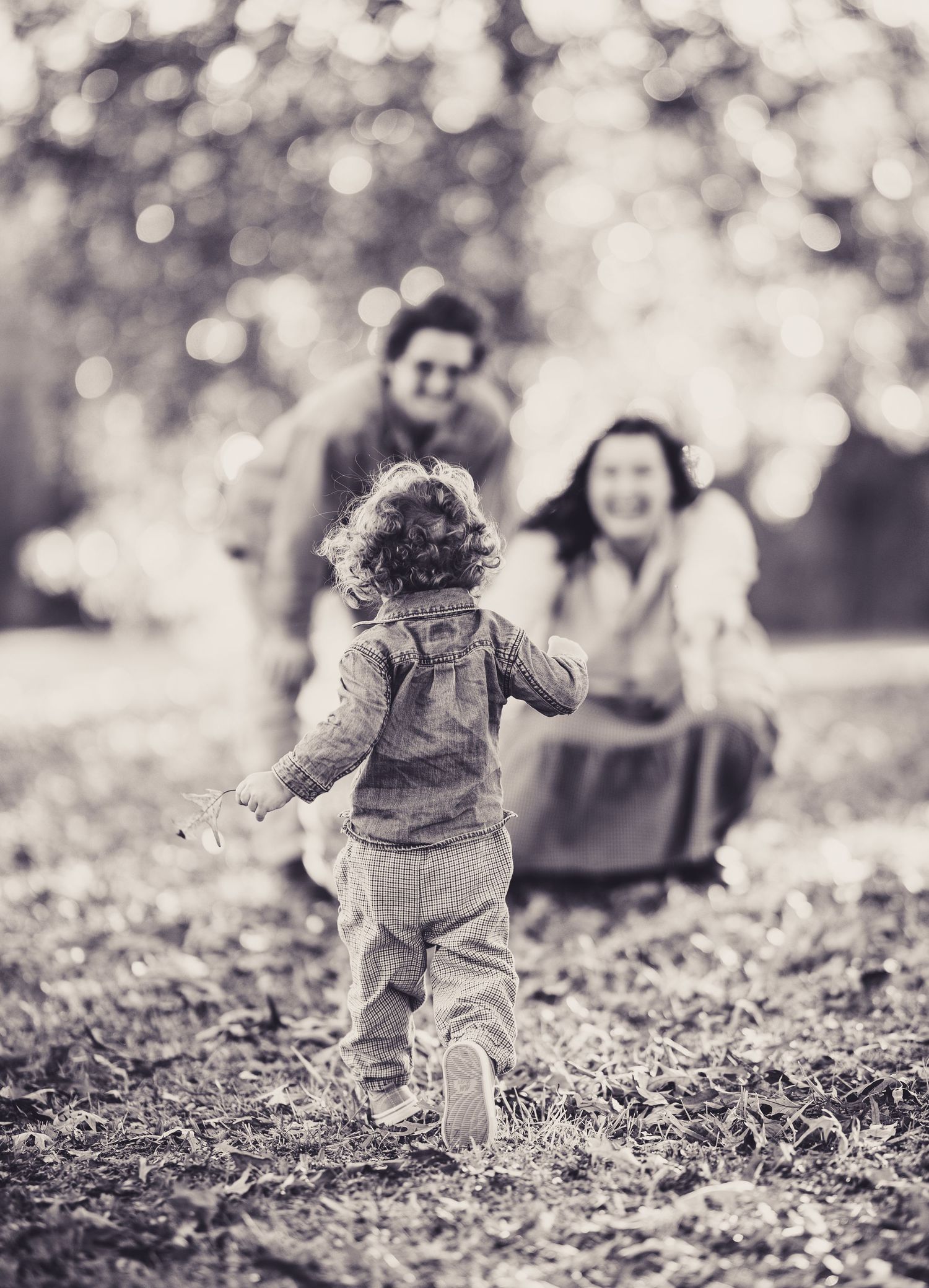 A toddler in overalls running towards adults on a fall day with bokeh background effects.