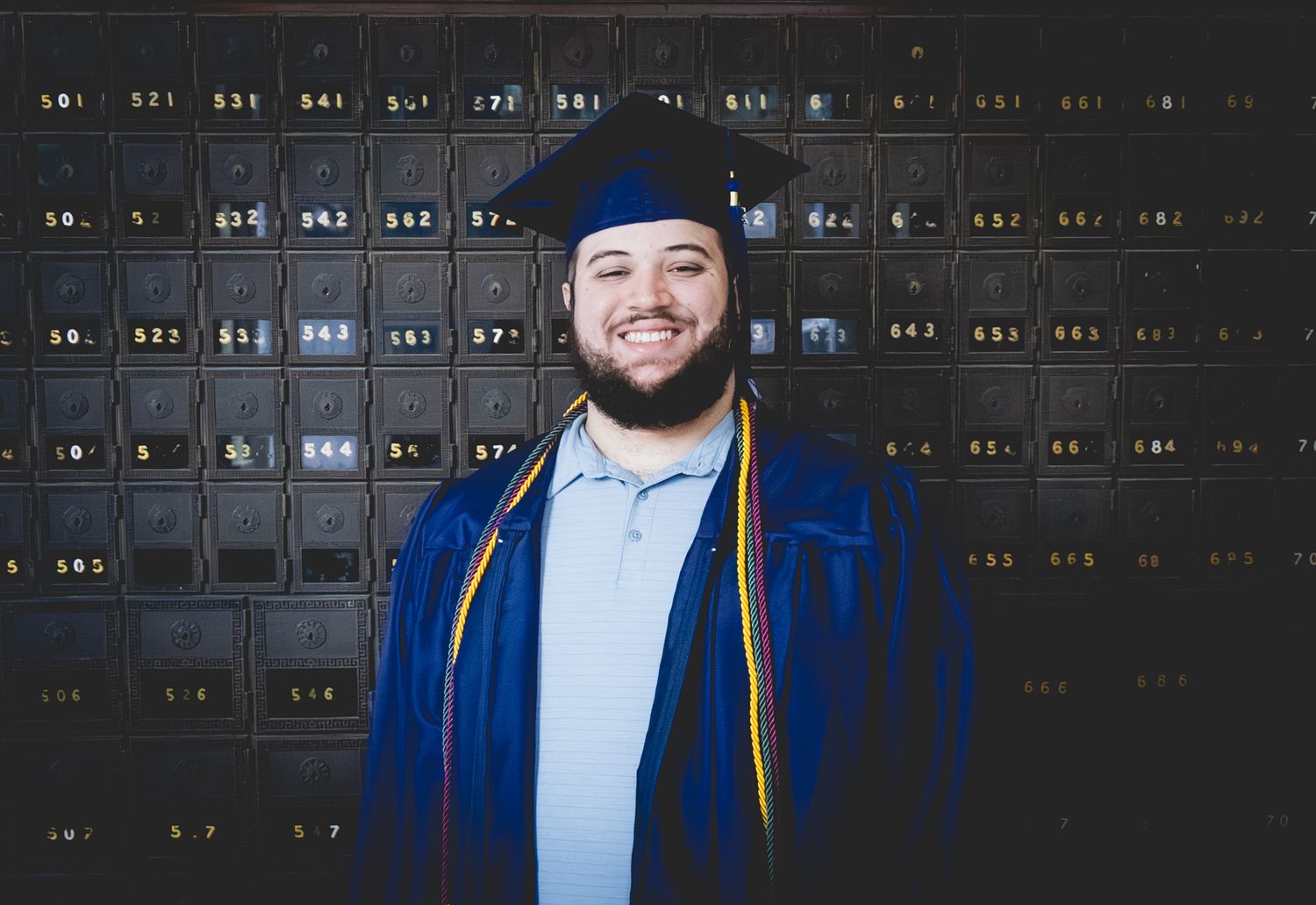 Smiling graduate in blue cap and gown stands proudly in front of dark lockers for a graduation portrait.