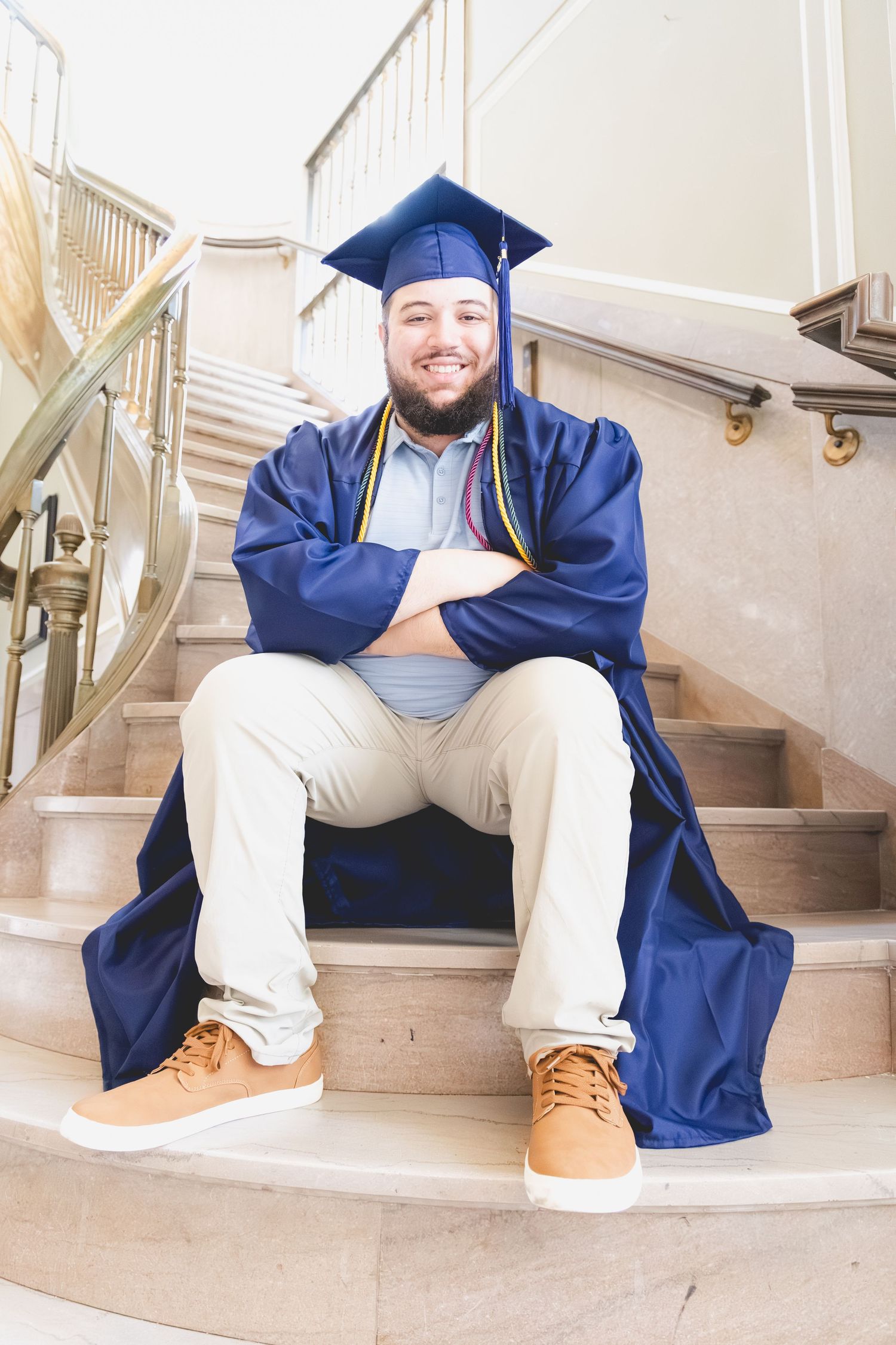 Smiling graduate in blue cap and gown sits on marble stairs with arms crossed, wearing khakis and tan sneakers.