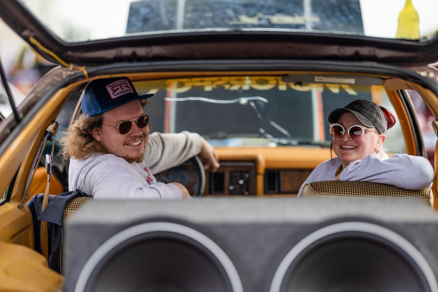 Two friends sitting in vintage car interior wearing sunglasses and casual attire.