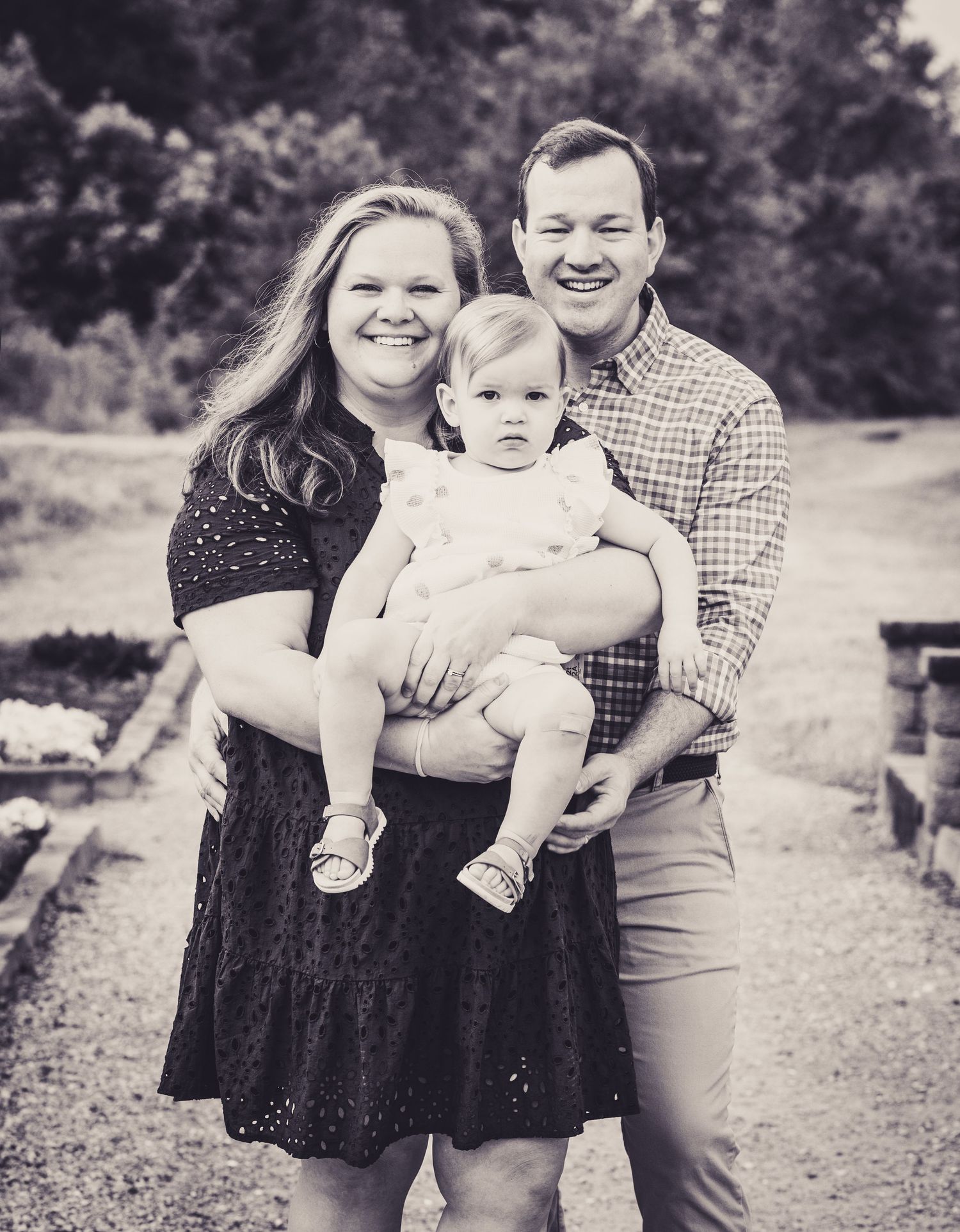 Black and white family portrait of smiling couple holding their baby girl outdoors on a garden path.