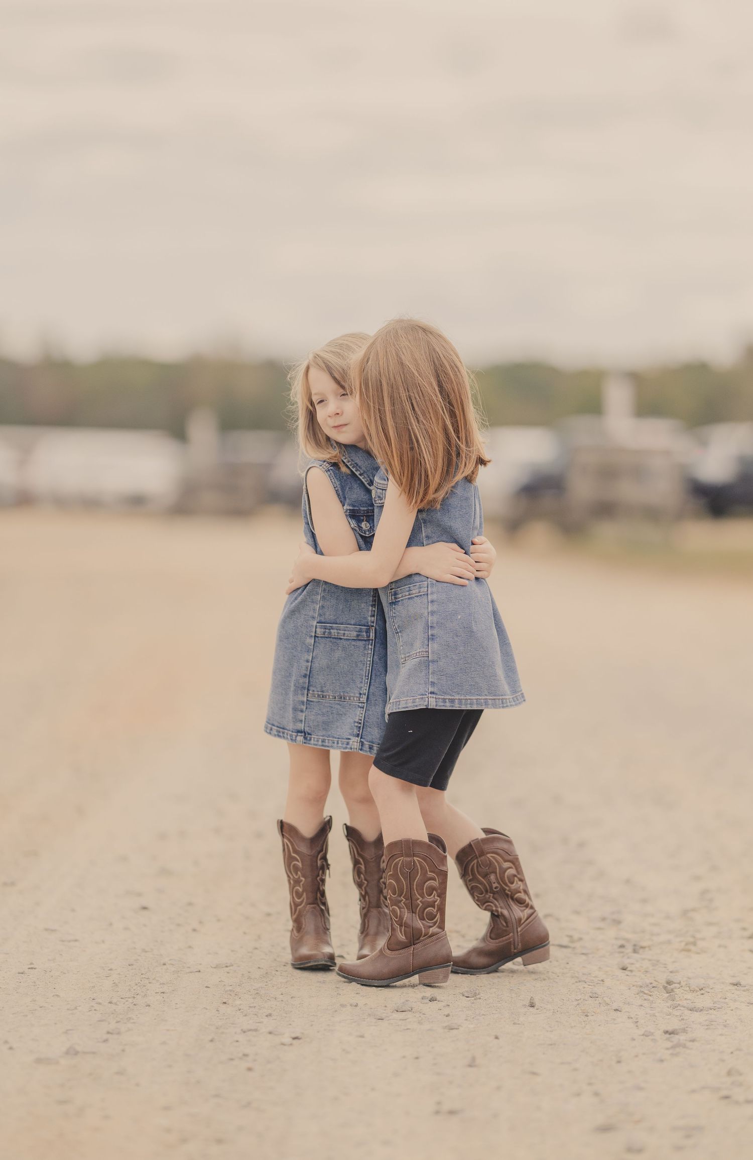 Two young girls in denim dresses and cowboy boots share a sweet hug on a dirt road.