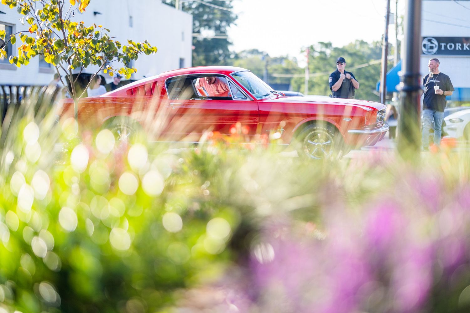 Classic red muscle car parked at a sunny outdoor car show surrounded by lush green and purple flowering plants.