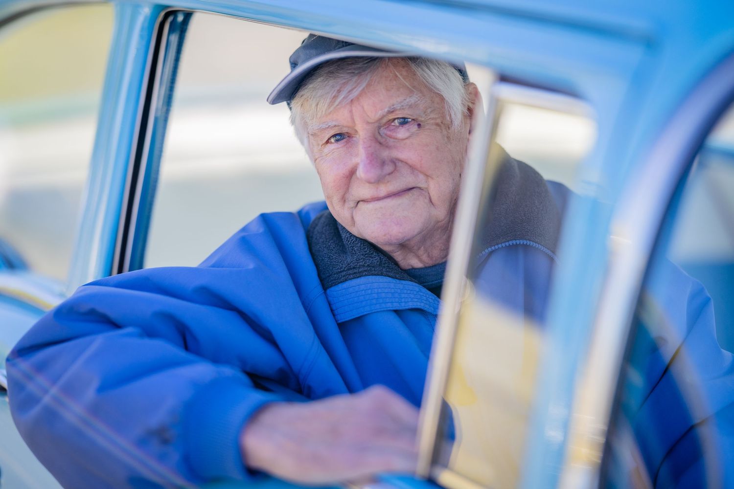 Elderly man in blue jacket and cap sitting in vintage blue car, smiling warmly through open window.