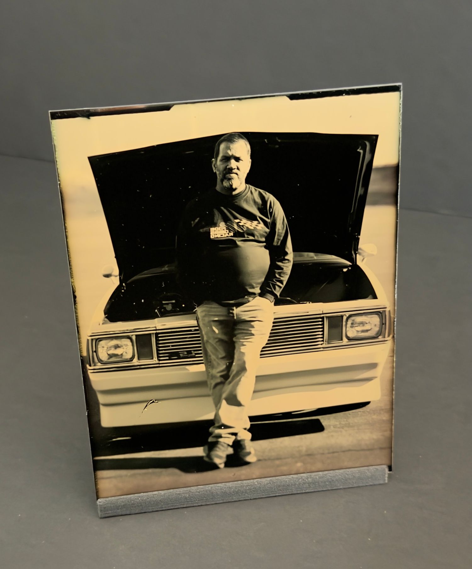Vintage black and white framed photo of a man leaning against a classic car with hood open.