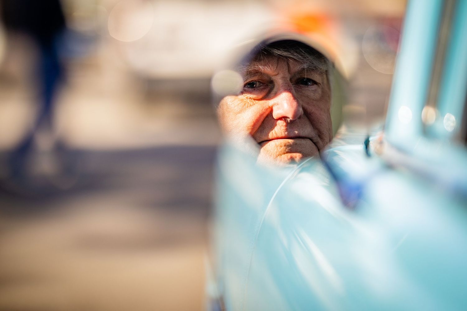 Elderly man's face reflected in the window of a vintage light blue car outdoors.