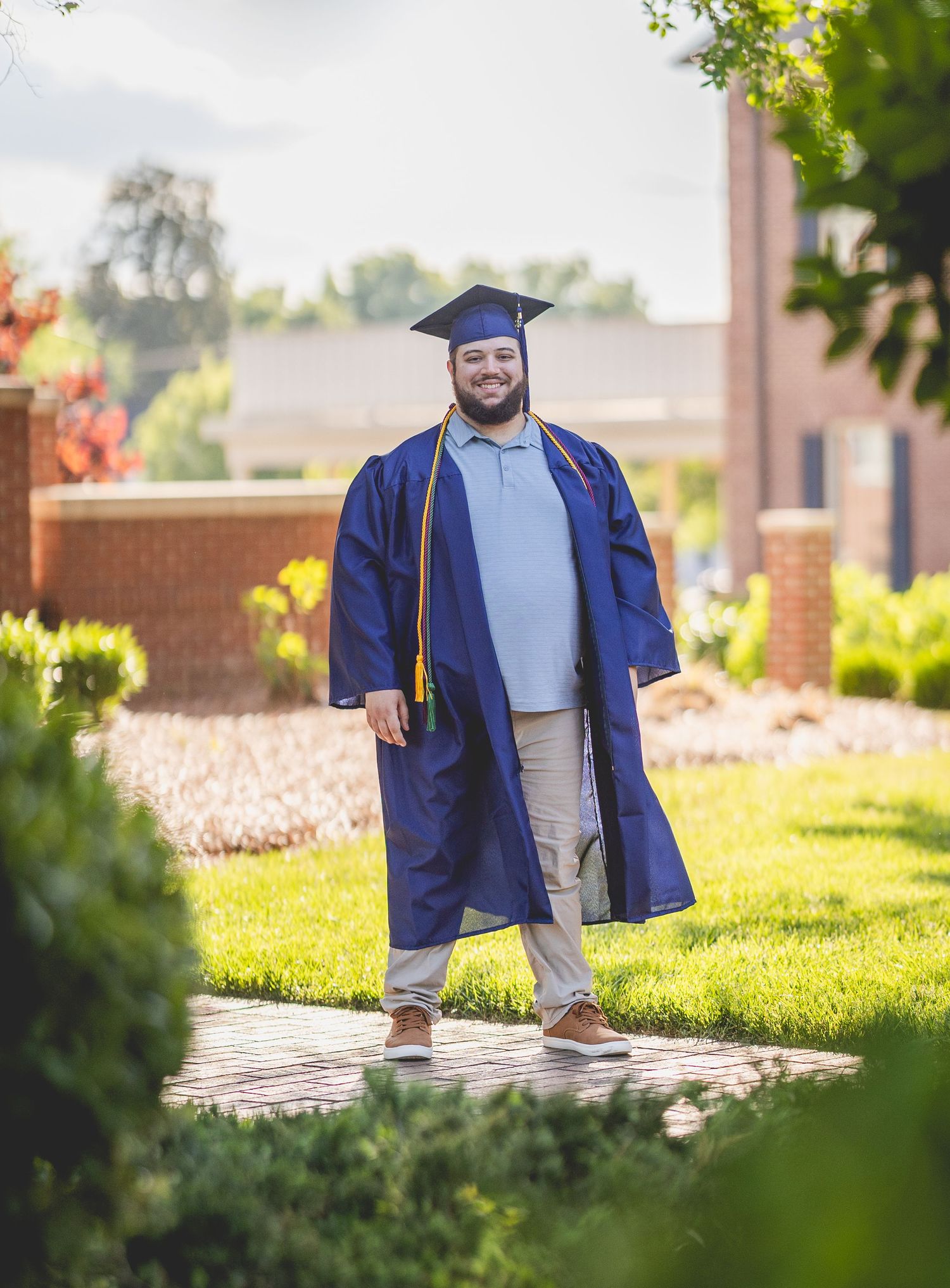 Smiling graduate in blue cap and gown walks along brick path on sunny college campus.