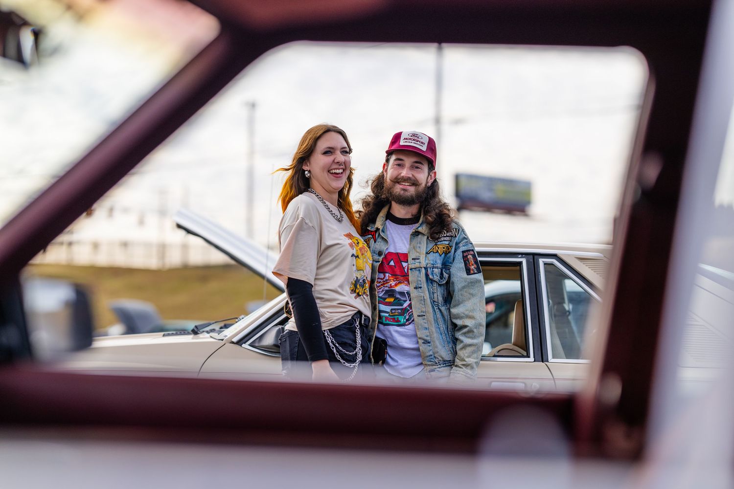 View through car window of couple standing by classic automobile at outdoor gathering.