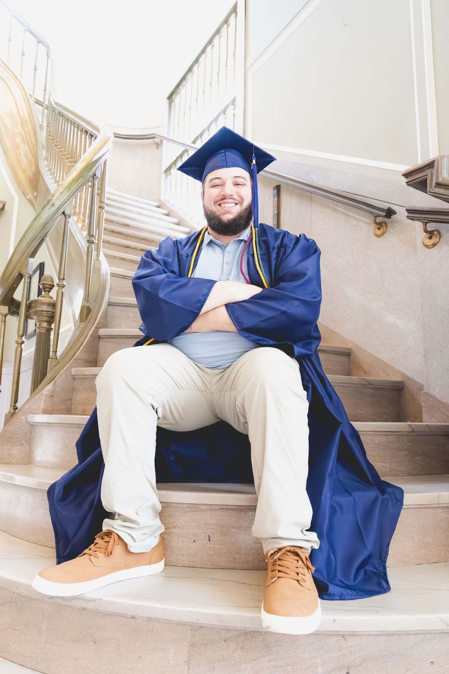 Smiling graduate in blue cap and gown sitting on marble stairs with arms crossed, wearing khakis and tan shoes.