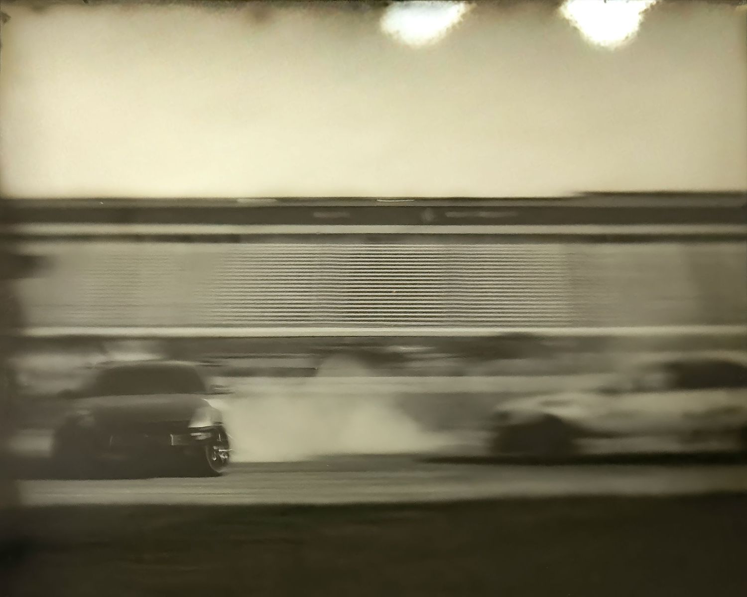 Blurry sepia-toned photo of racing cars drifting on a track, smoke billowing, captured with motion blur effect.