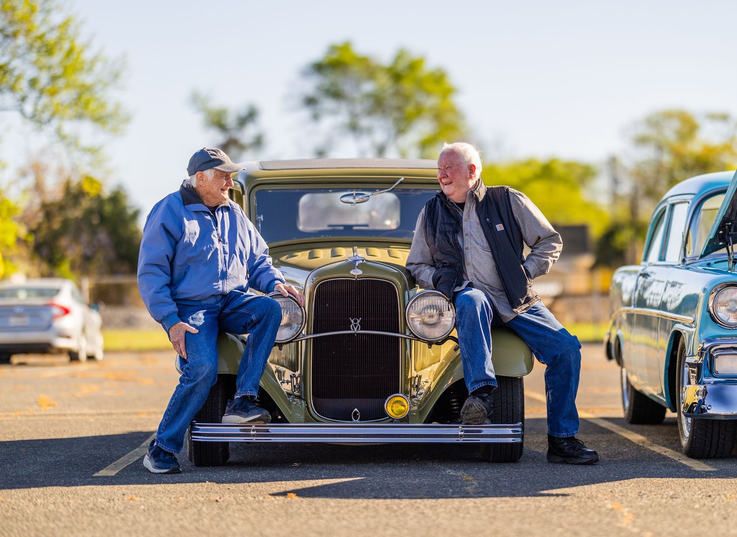 Two men sit on the hood of a vintage classic car at an outdoor car show on a sunny day.