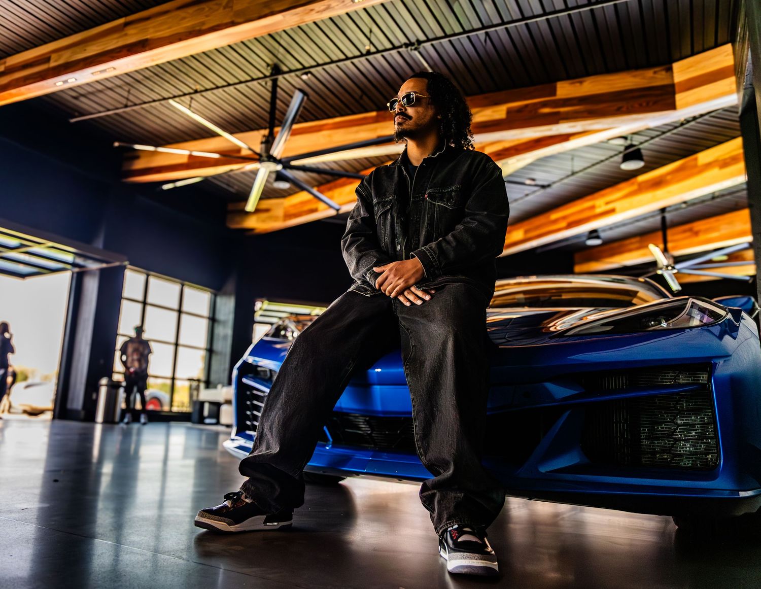 Man in black jacket leaning on blue sports car in modern showroom with wood ceiling.