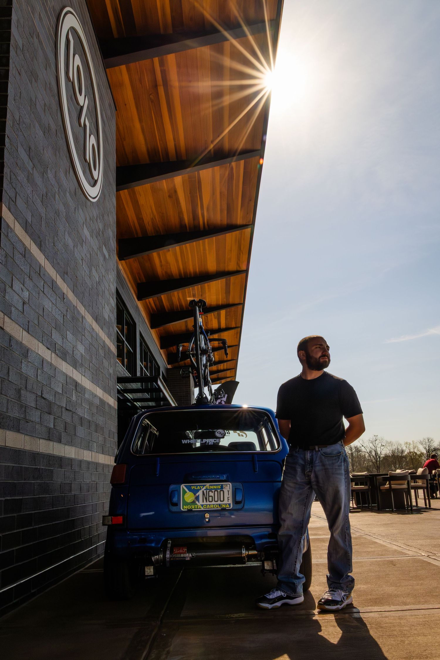 Man standing beside a blue SUV with bike rack outside a brick building, sun flaring dramatically overhead.