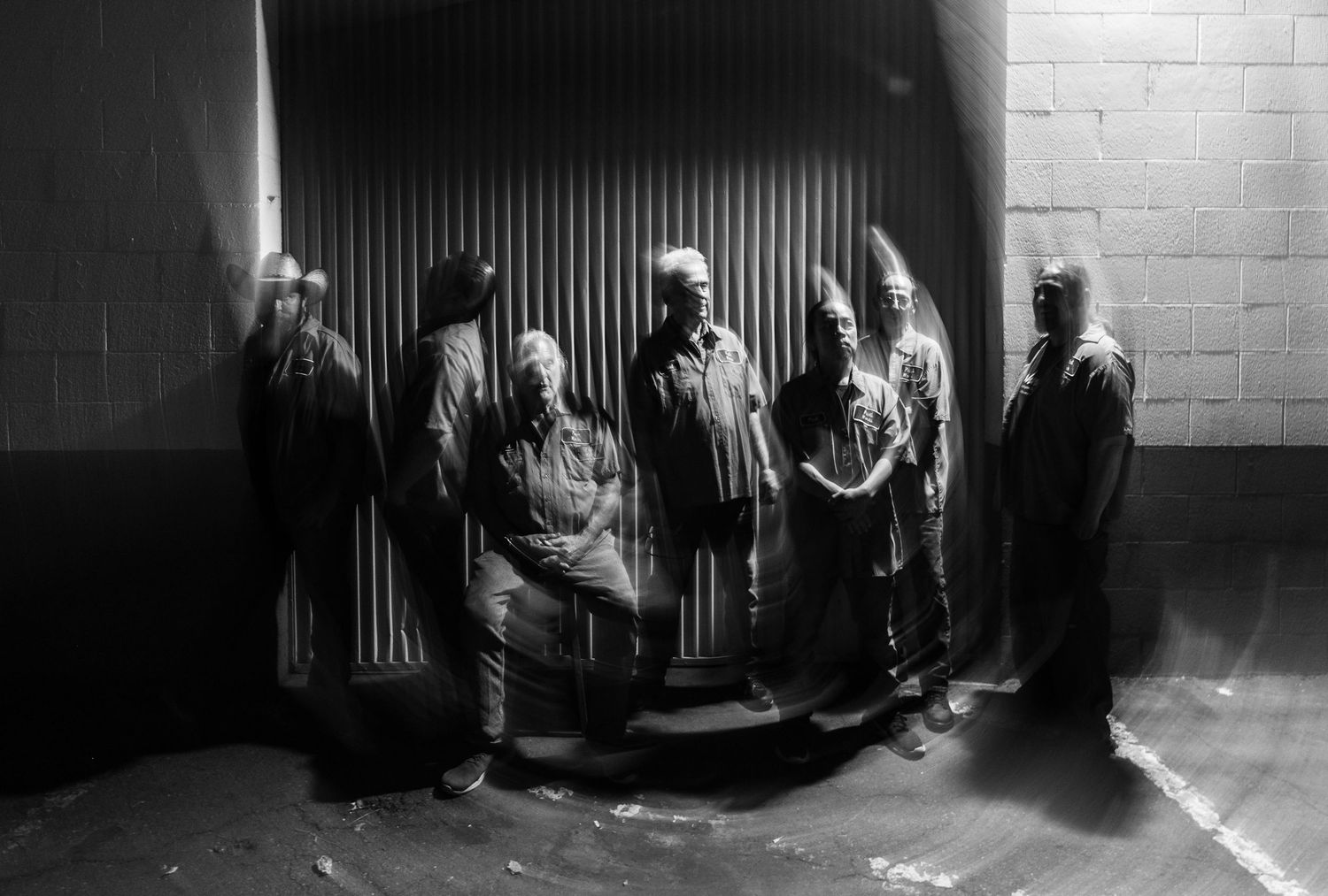 A dramatic black and white photo of people standing against a wall in dark clothing.