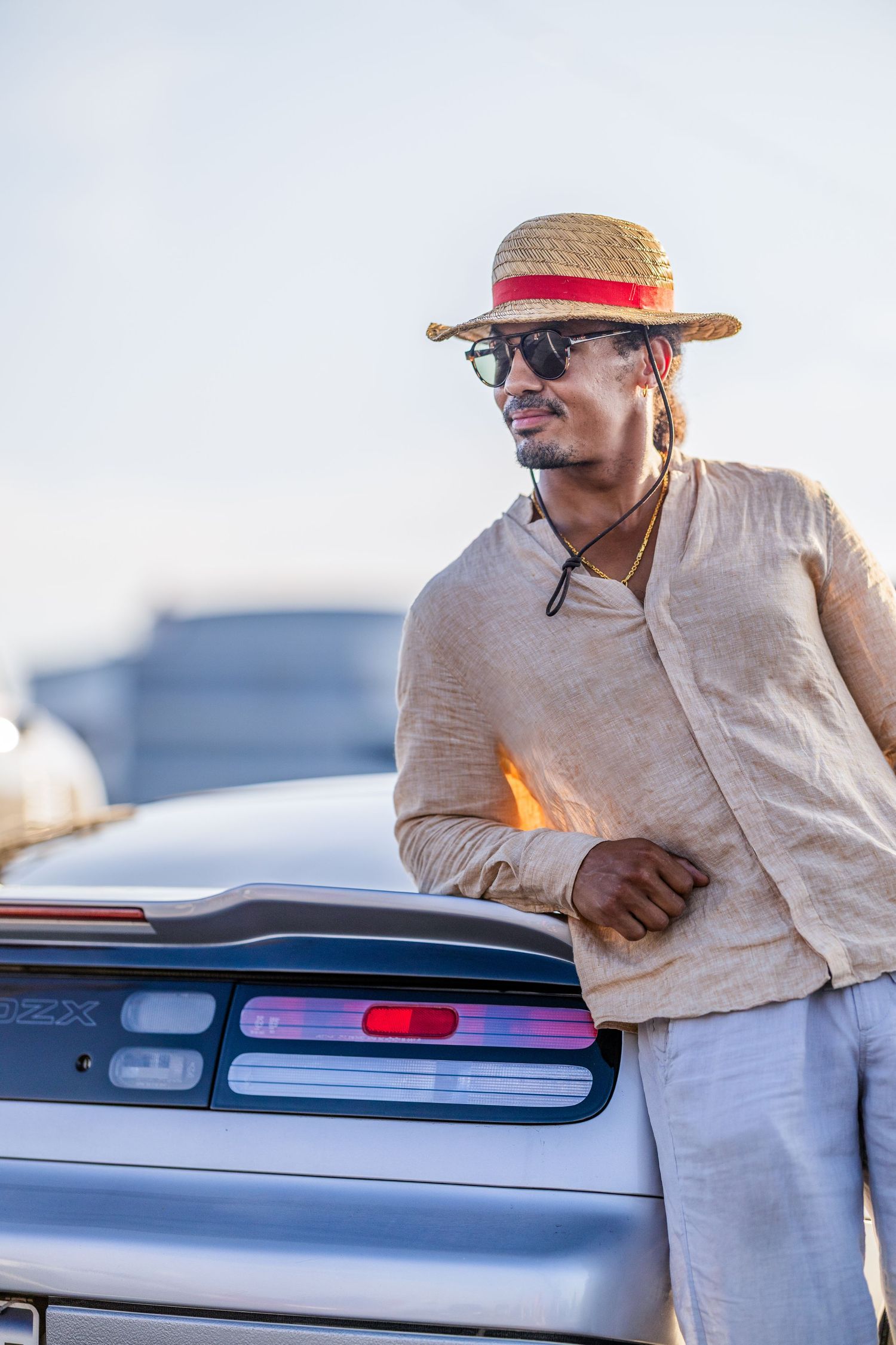 Person wearing a straw hat and light colored clothing leans against a classic car at a show.