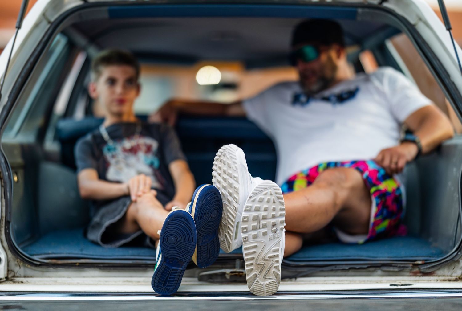 Man and child relaxing in open car trunk with feet up, enjoying a casual road trip together on a sunny day.