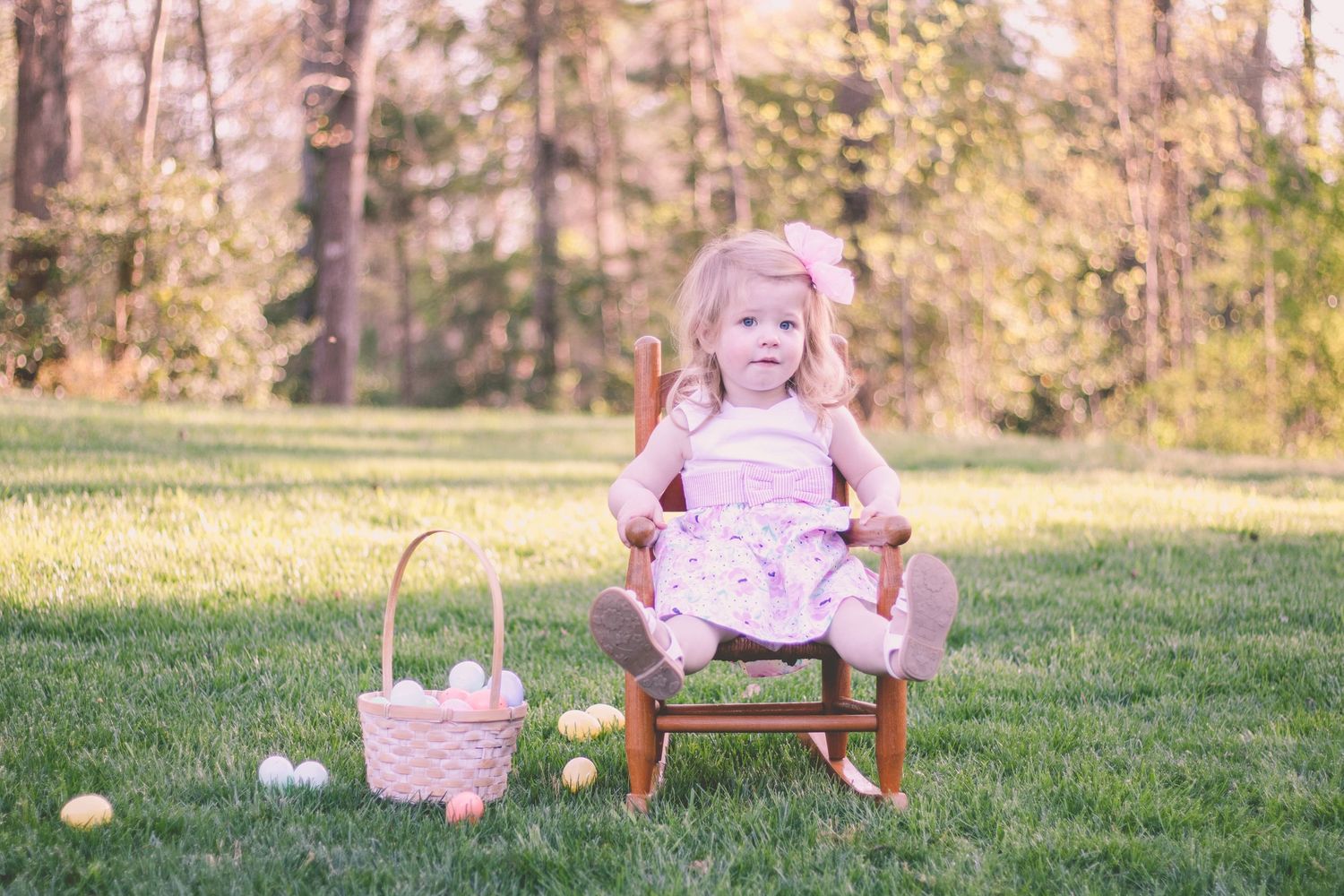 Toddler girl in pink dress sitting on wooden chair outdoors with Easter basket and eggs on green grass.