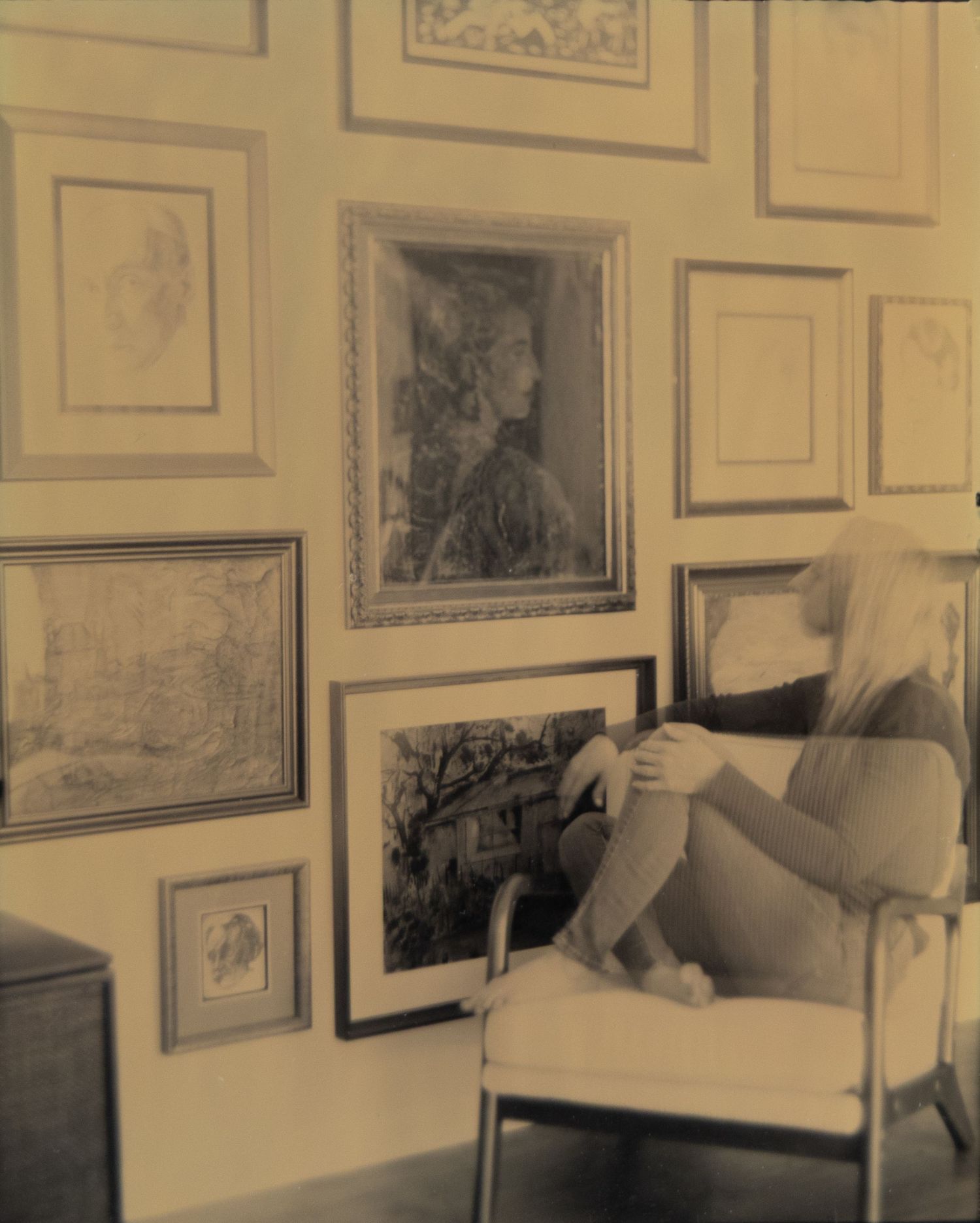 Vintage sepia photo of a woman relaxing in a chair surrounded by framed artwork and sketches on a gallery wall.