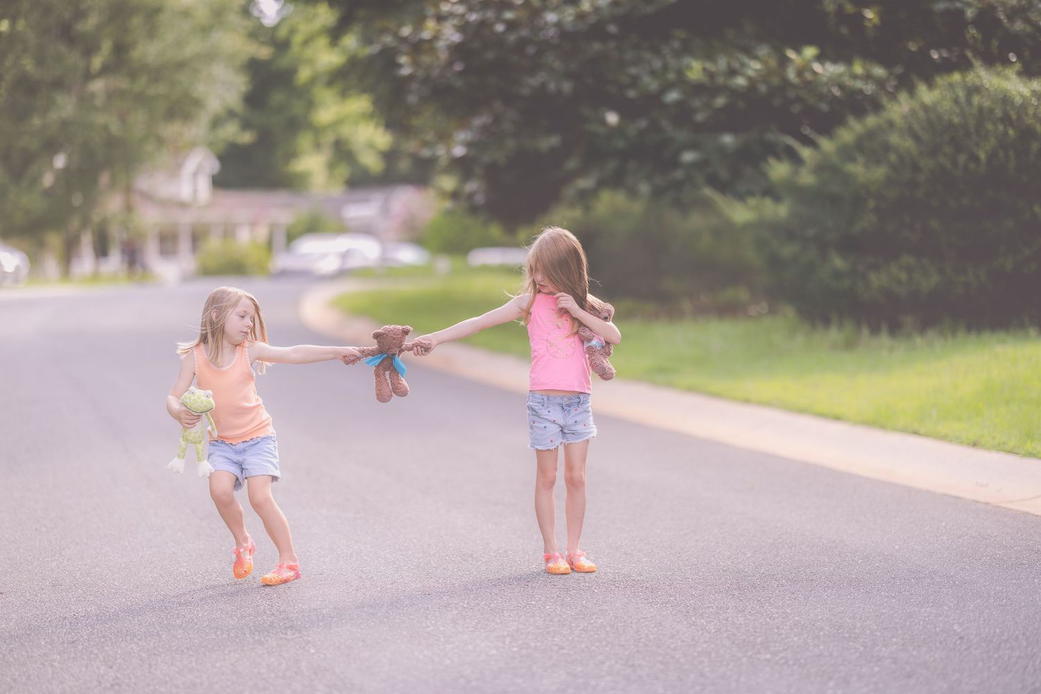 Two young girls play tug-of-war with a teddy bear on a sunny suburban street.