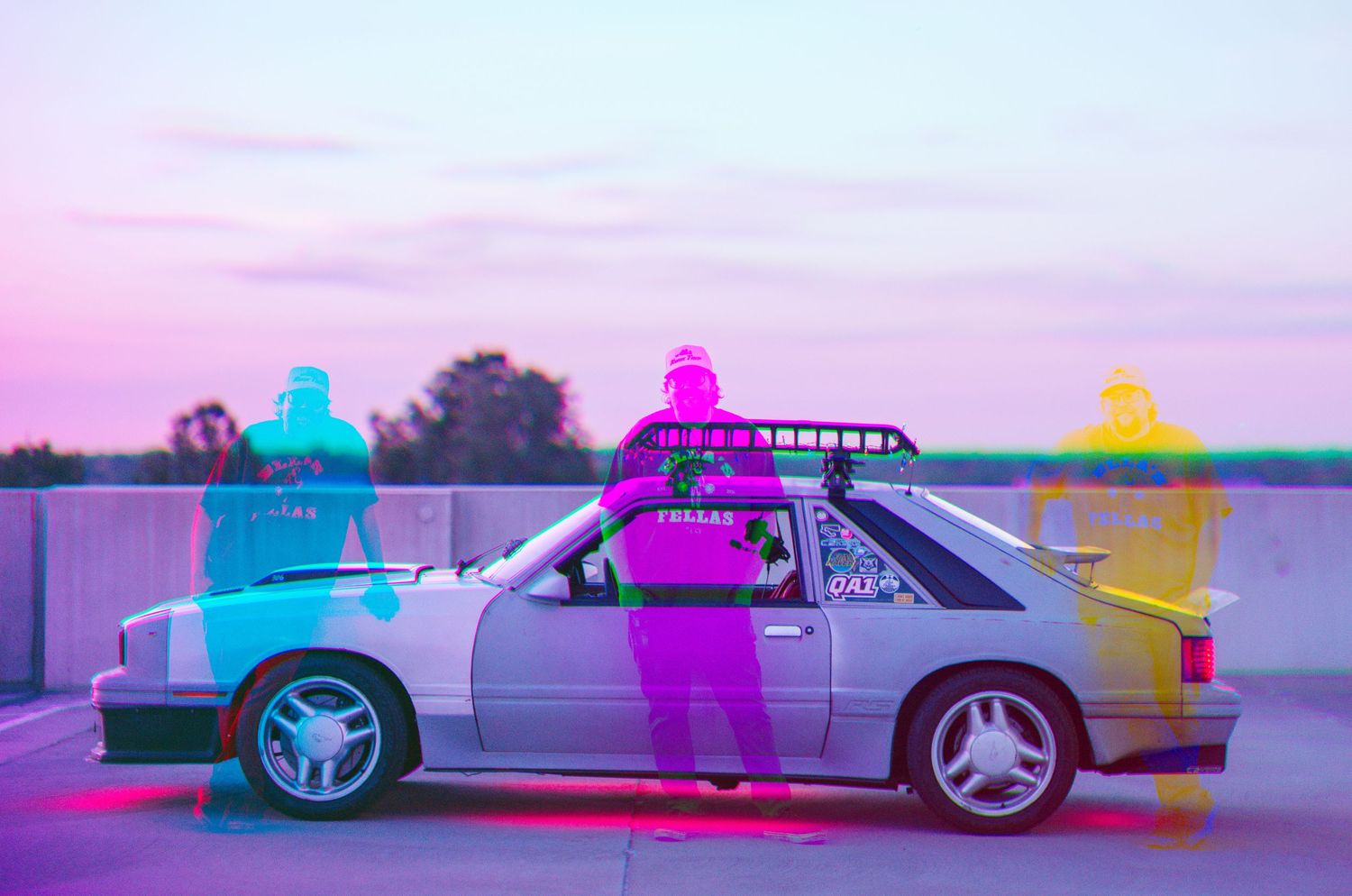 A silver car on a rooftop parking lot with colorful neon ghost-like figures in pink, cyan, and yellow at sunset.