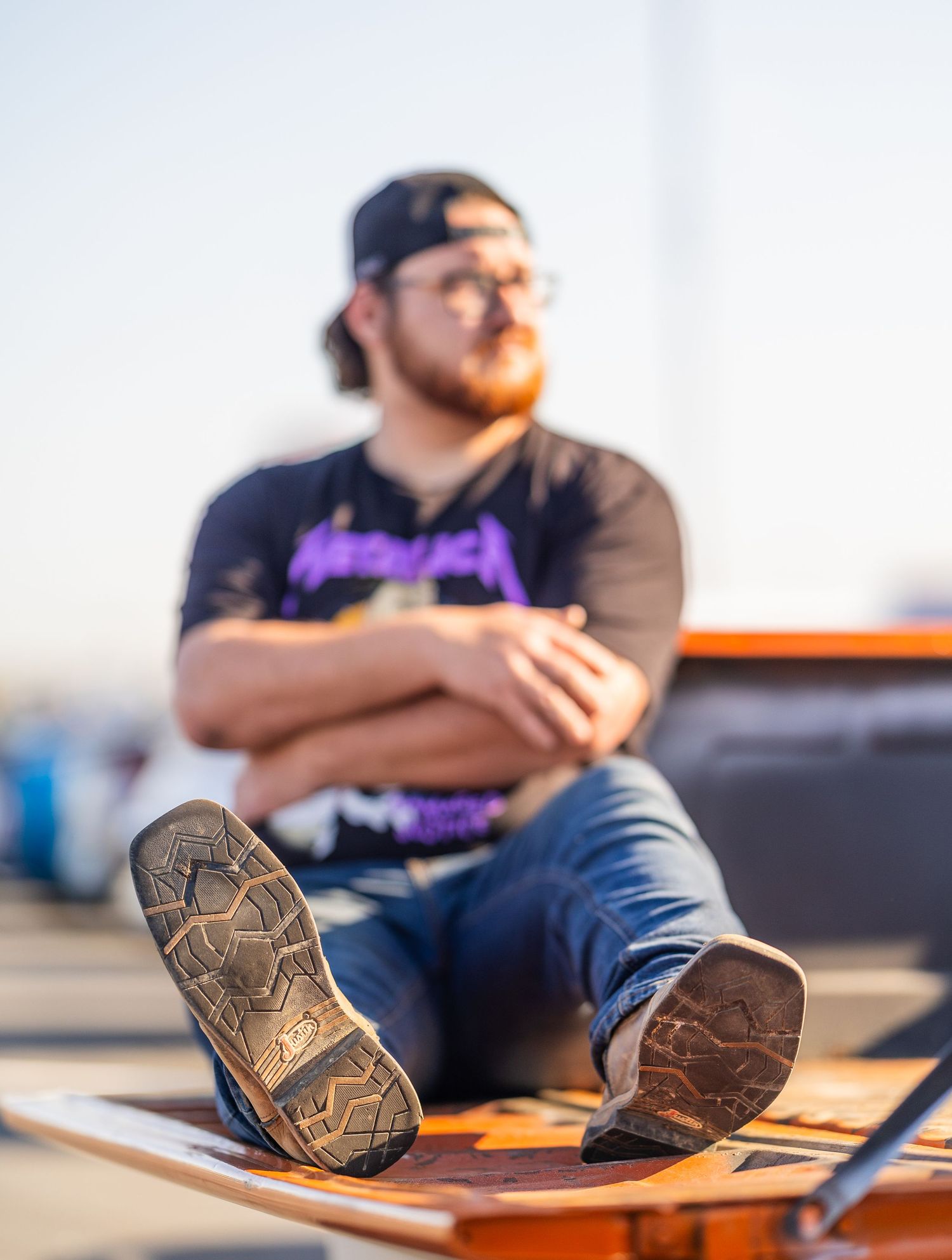 Man in Metallica shirt and cap sits relaxed outdoors, boots in foreground with shallow depth of field bokeh effect.