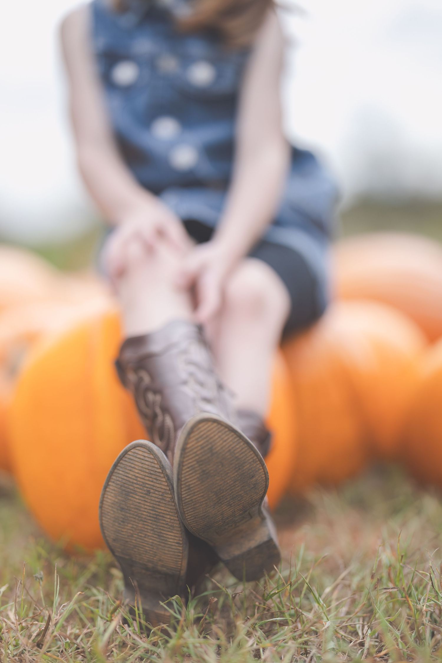 Young girl in denim dress sitting on orange pumpkins showing soles of brown cowboy boots in a fall field.