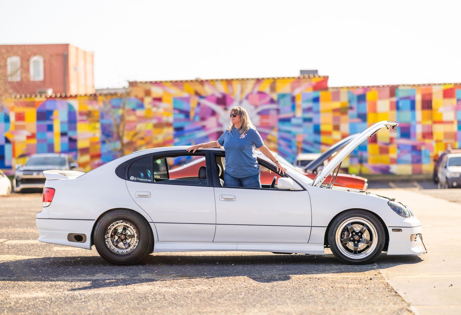 A white sports car with custom modifications parked in front of a colorful wall mural.